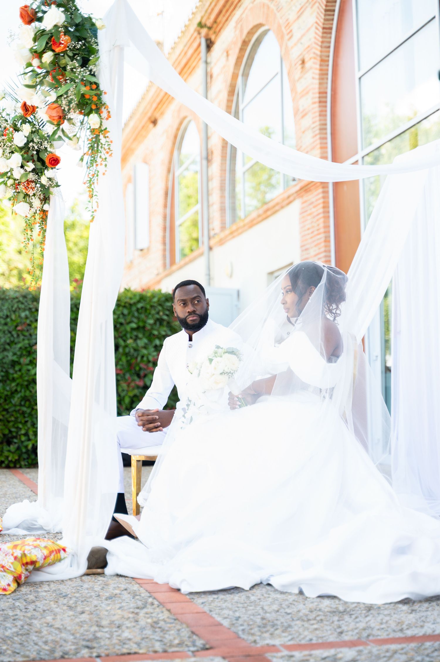 Elegant bride and groom seated under white draped wedding canopy with orange floral decor.