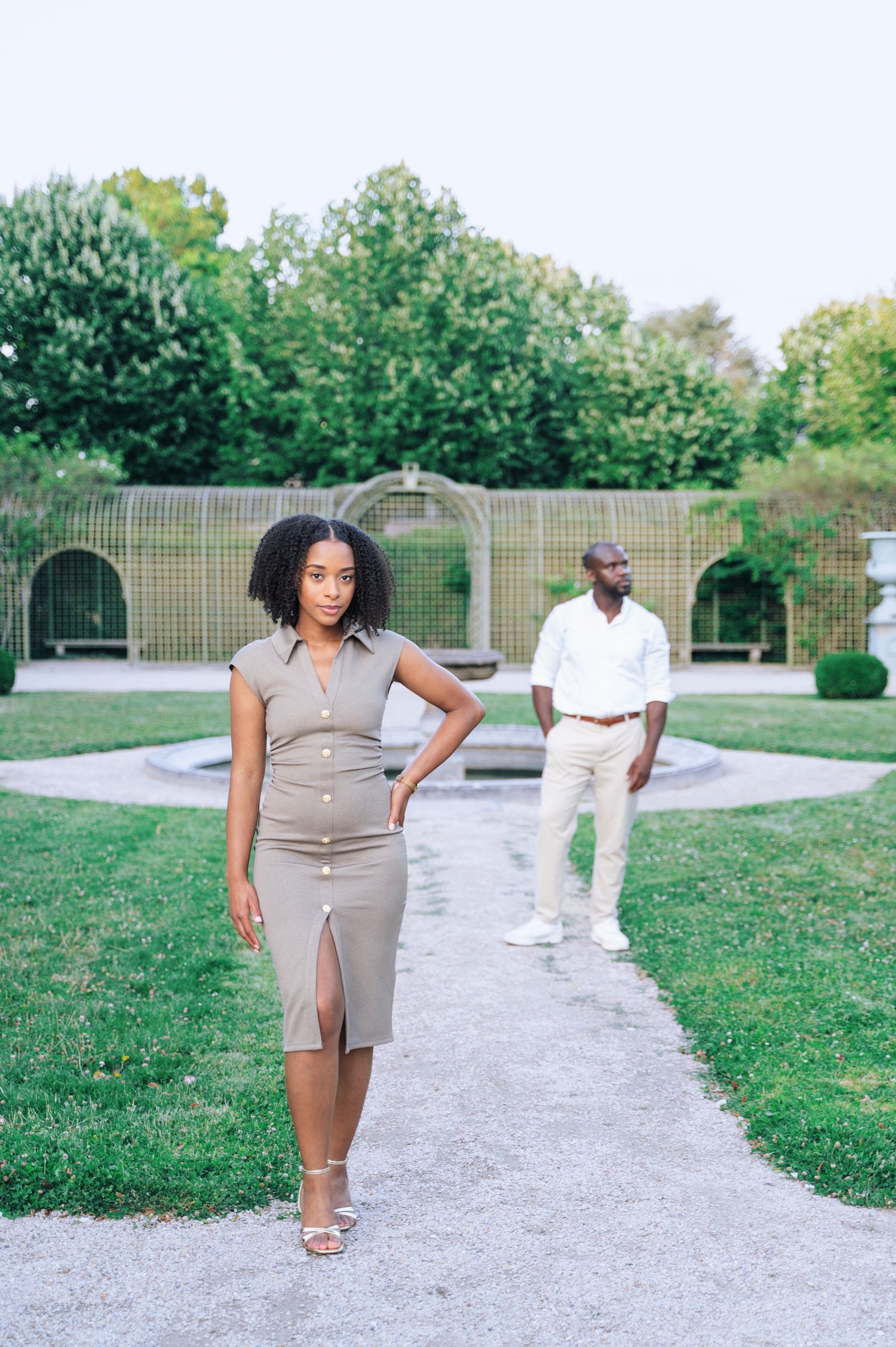 A young Black woman in a taupe dress stands confidently in a lush garden with a man in white standing behind her.