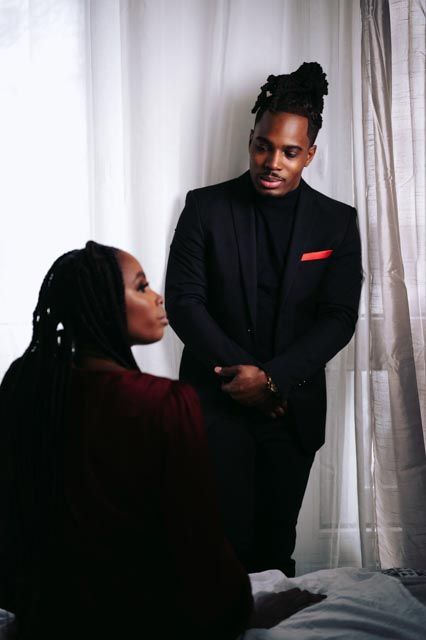 A man in a black suit with red pocket square holds hands with a seated woman with braids near a bright window.