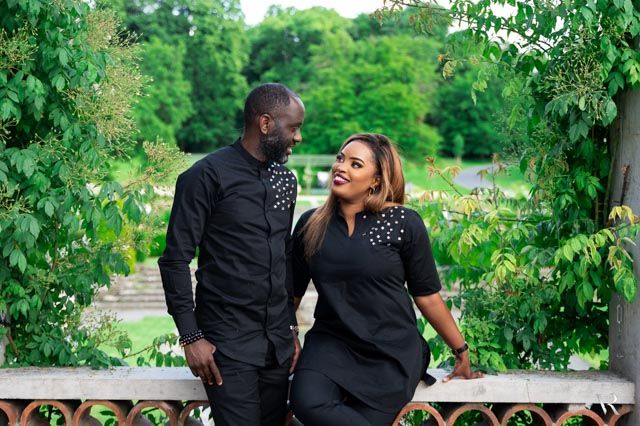 Couple in matching black outfits smiling at each other in a lush green garden park setting.