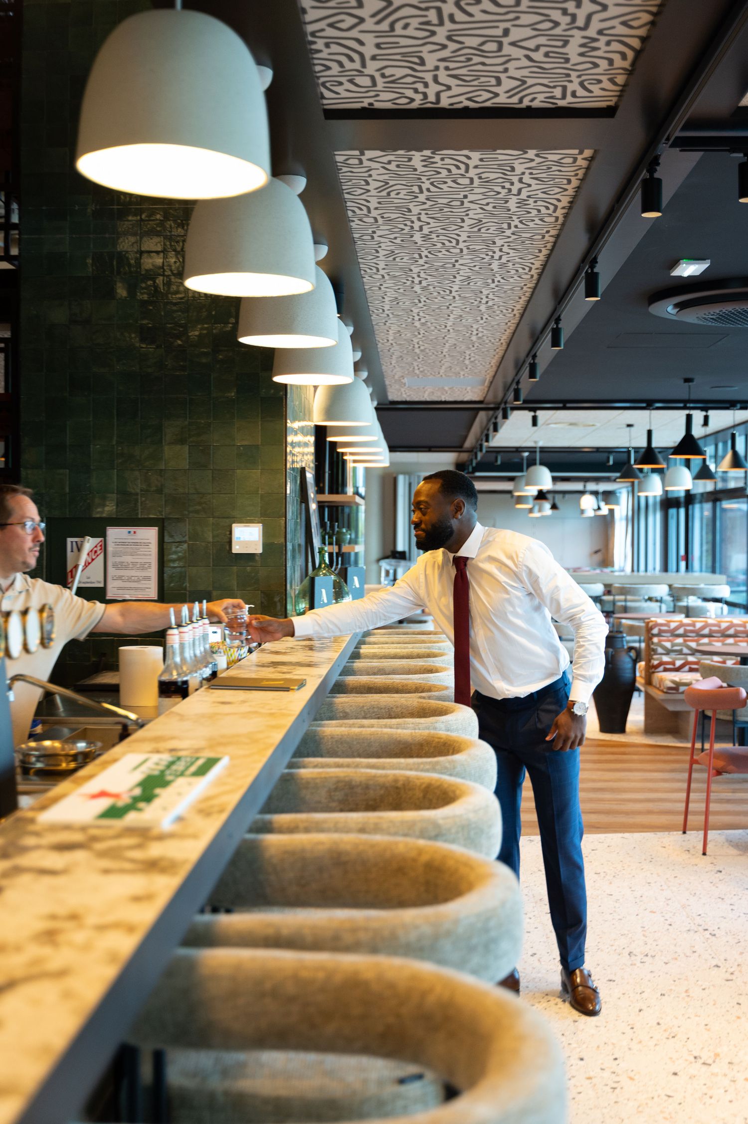 Dapper Black man in white shirt and red tie interacts with staff at a sleek modern restaurant bar.