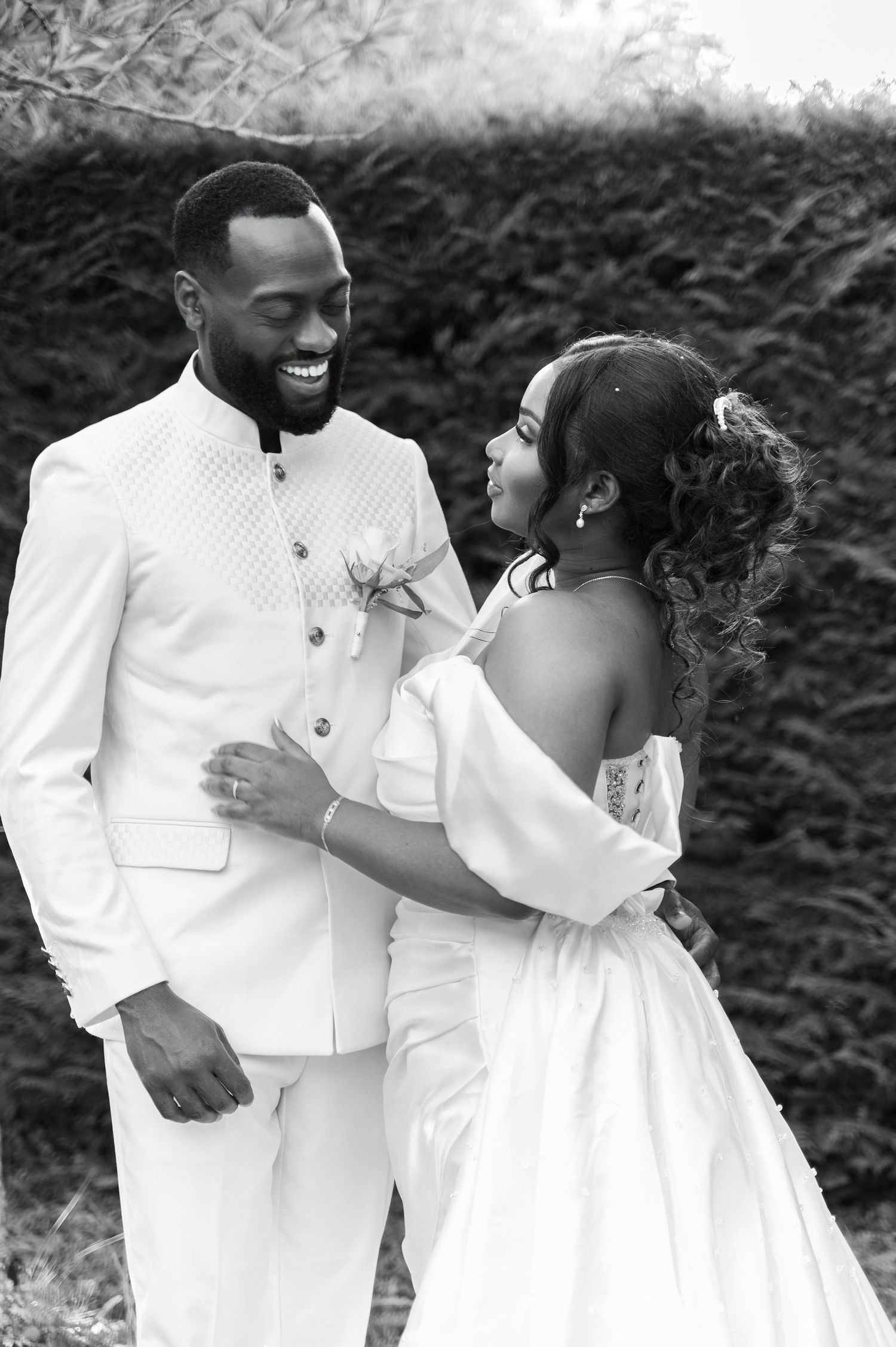 Black and white photo of a smiling groom in white suit embracing his bride in an off-shoulder wedding gown outdoors.