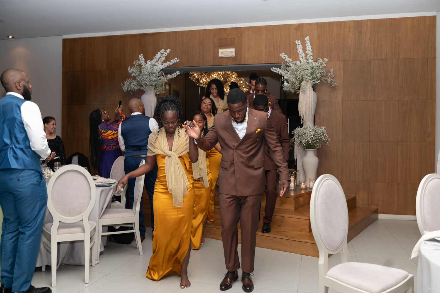 Groom in brown suit escorts a woman in golden dress into a modern wedding ceremony venue.