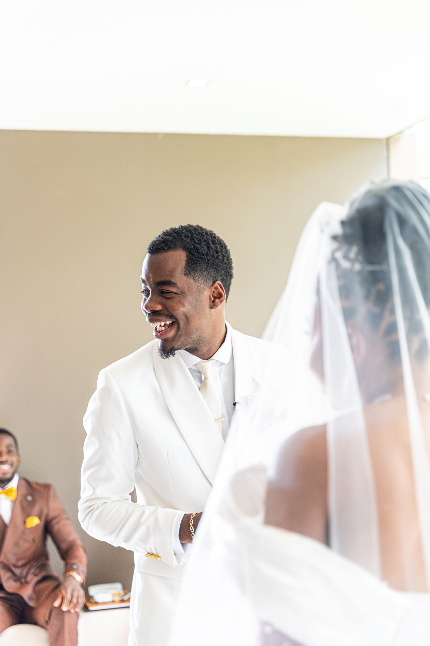 Smiling groom in white tuxedo faces bride in veil during intimate wedding ceremony moment.
