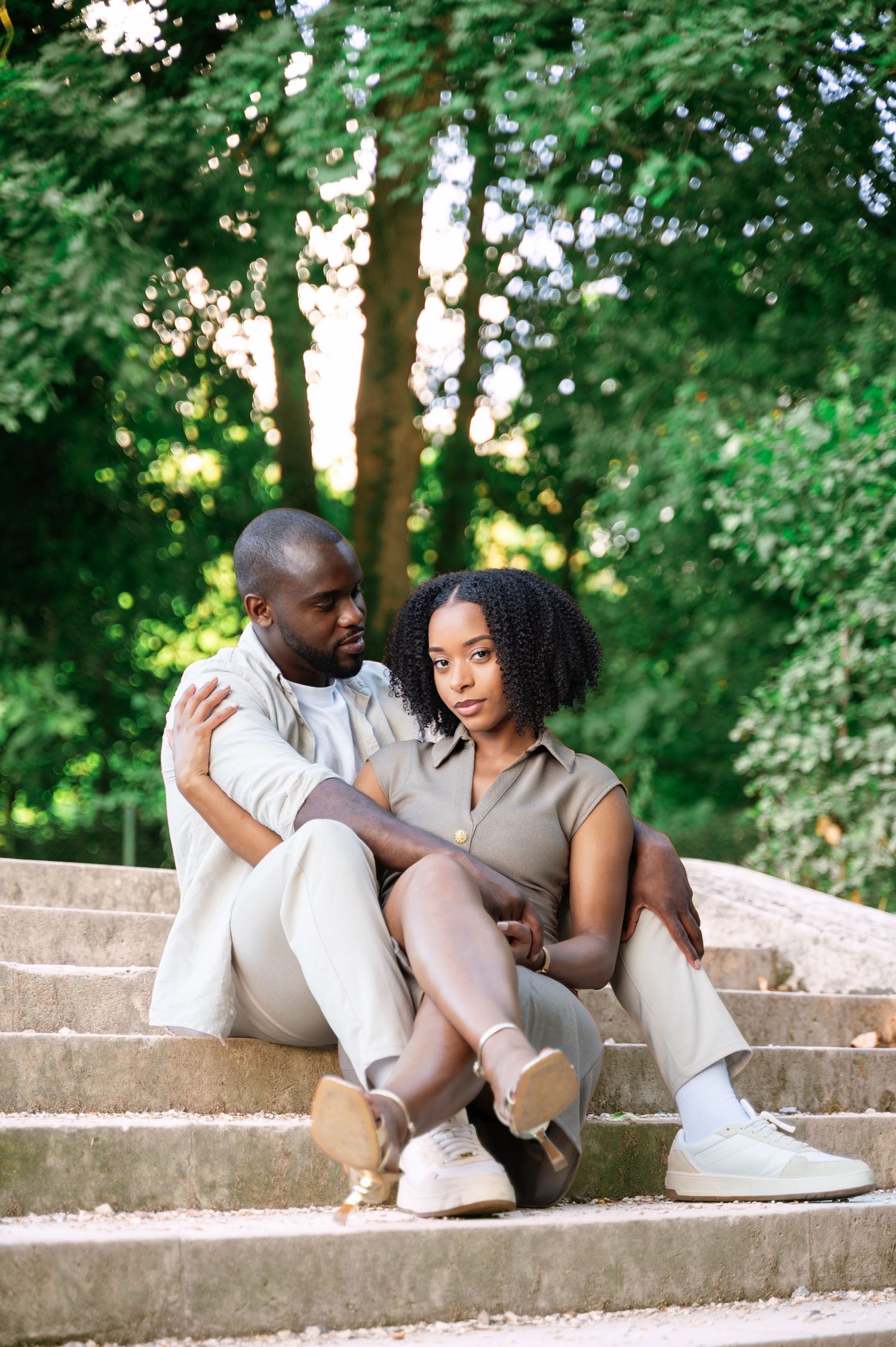 A stylish Black couple sits relaxed on outdoor stone steps surrounded by vibrant green trees in a park.