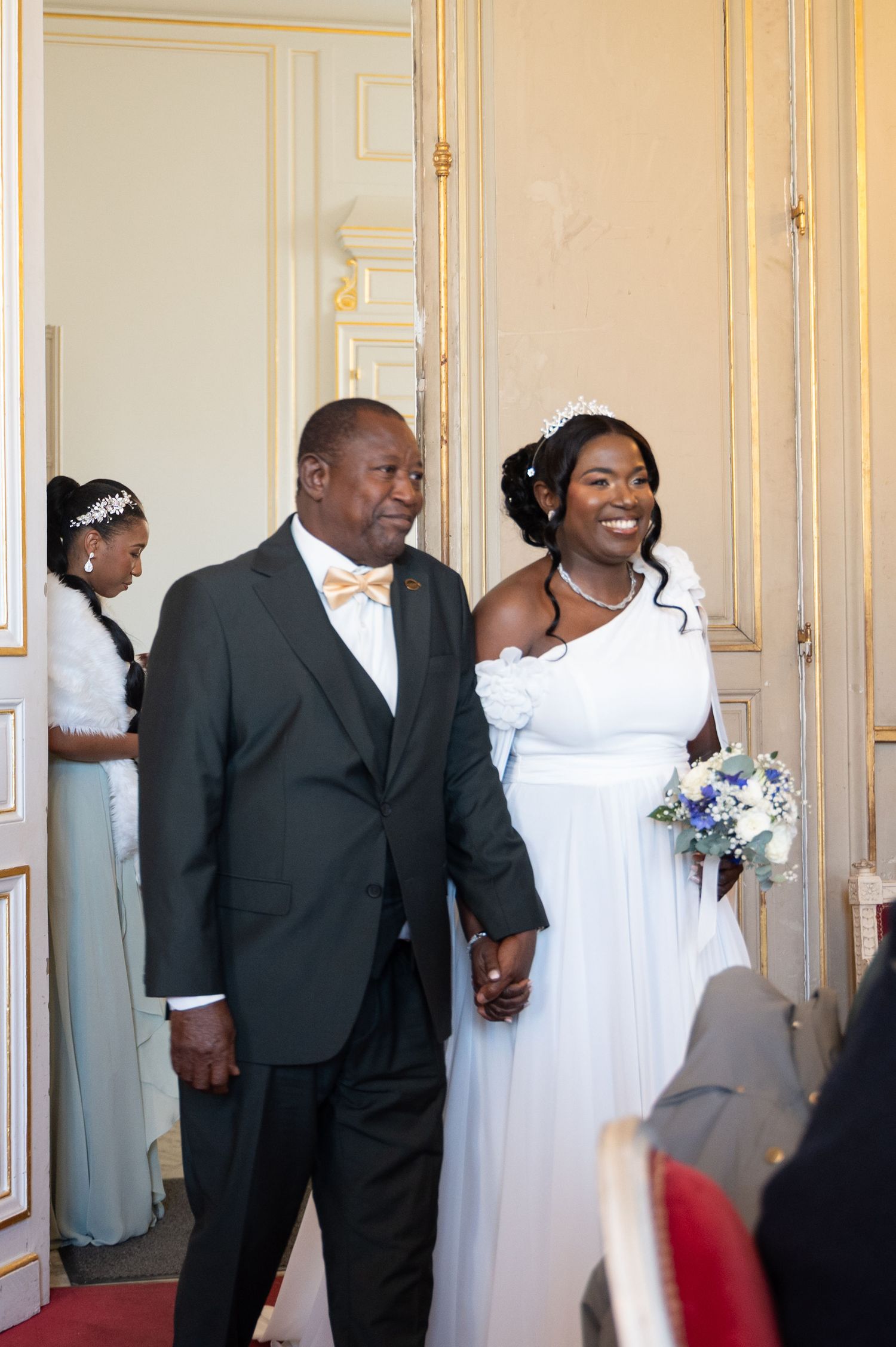 A radiant Black bride in a white off-shoulder gown walks the aisle holding hands with an older man in a dark suit.