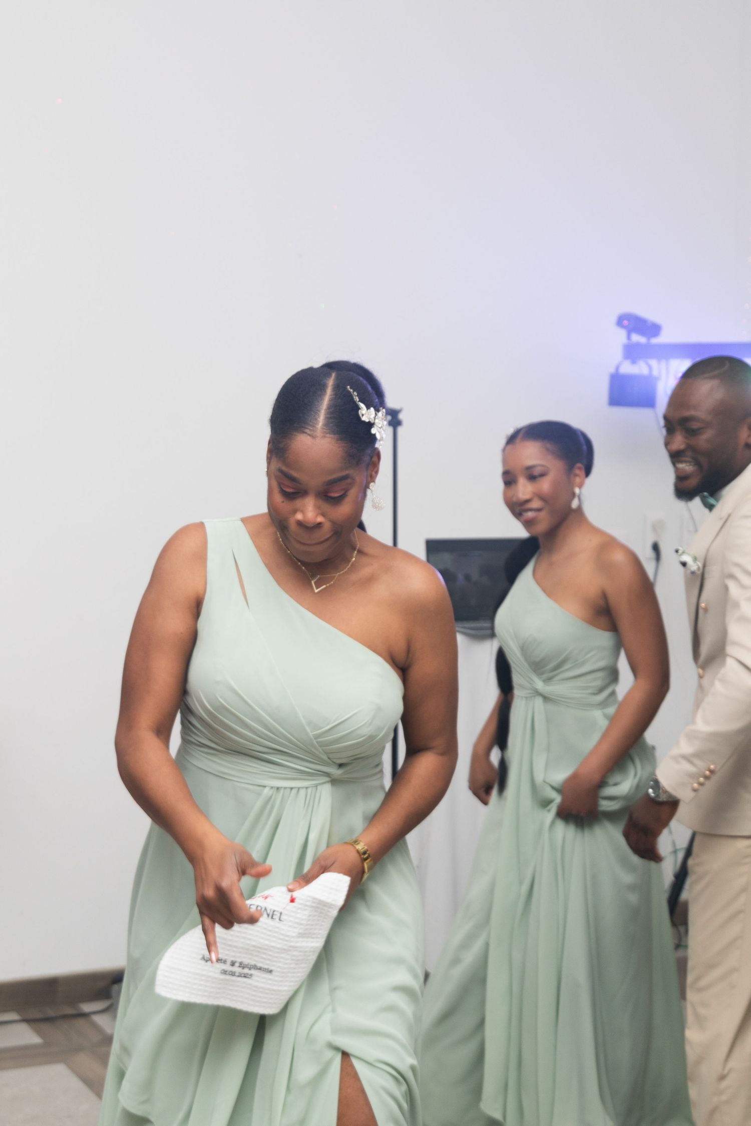 Bridesmaids in sage green one-shoulder gowns walk together at an elegant wedding reception event.