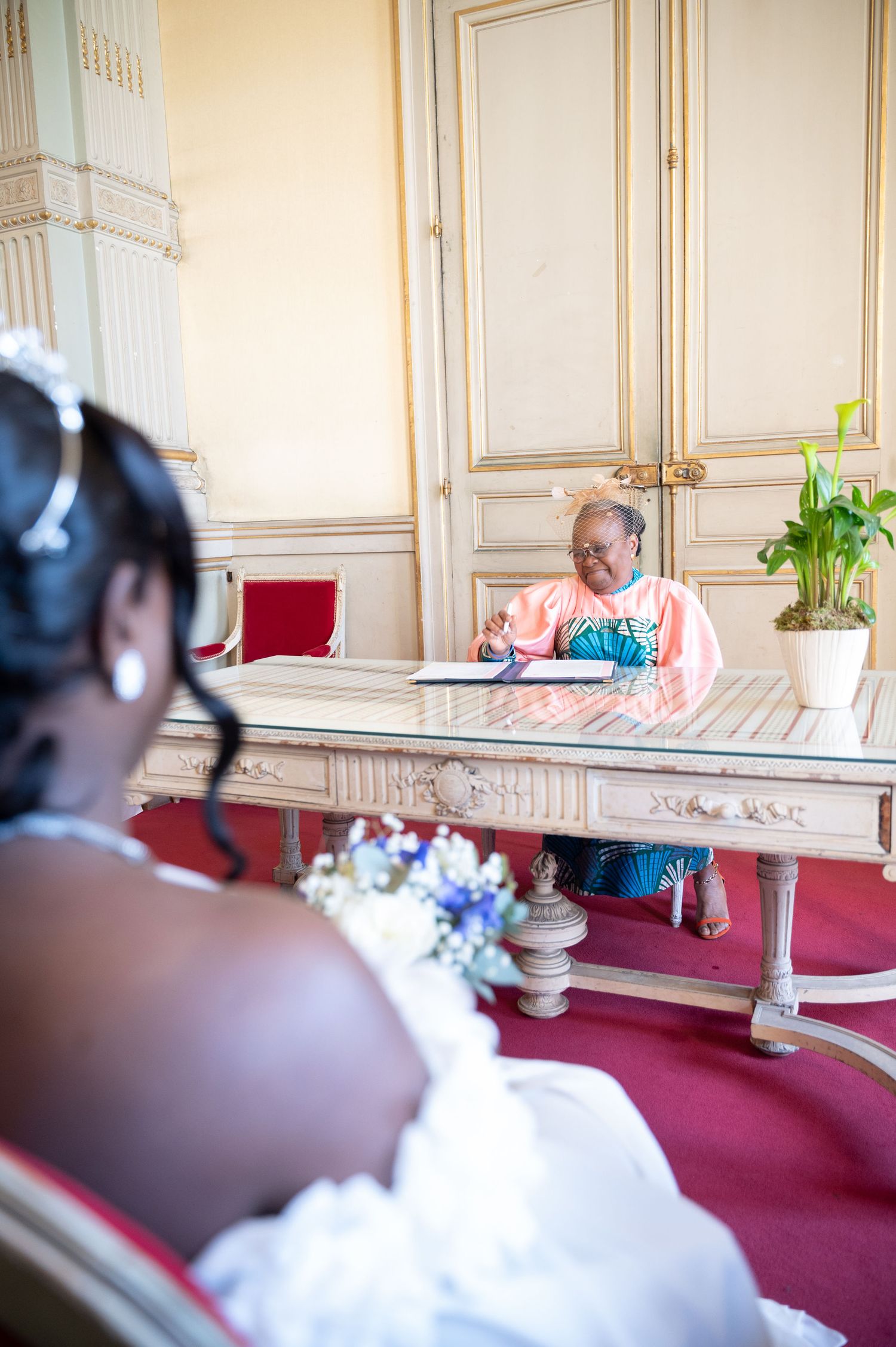 A Black bride in a white gown with tiara sits before an official signing a document at an ornate wedding registry desk.