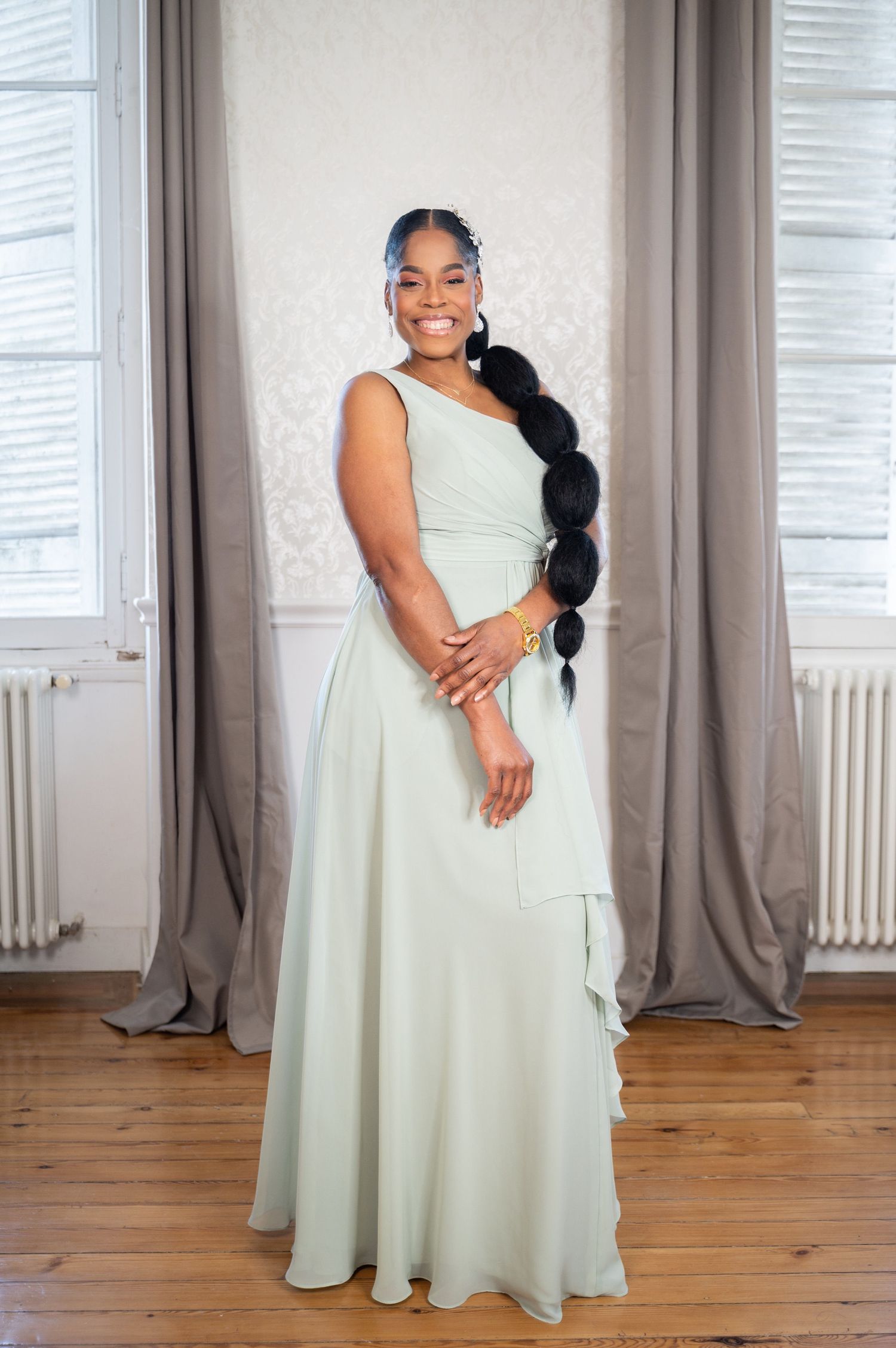 Woman in elegant mint green one-shoulder gown with long braided hair posing in a bright room.