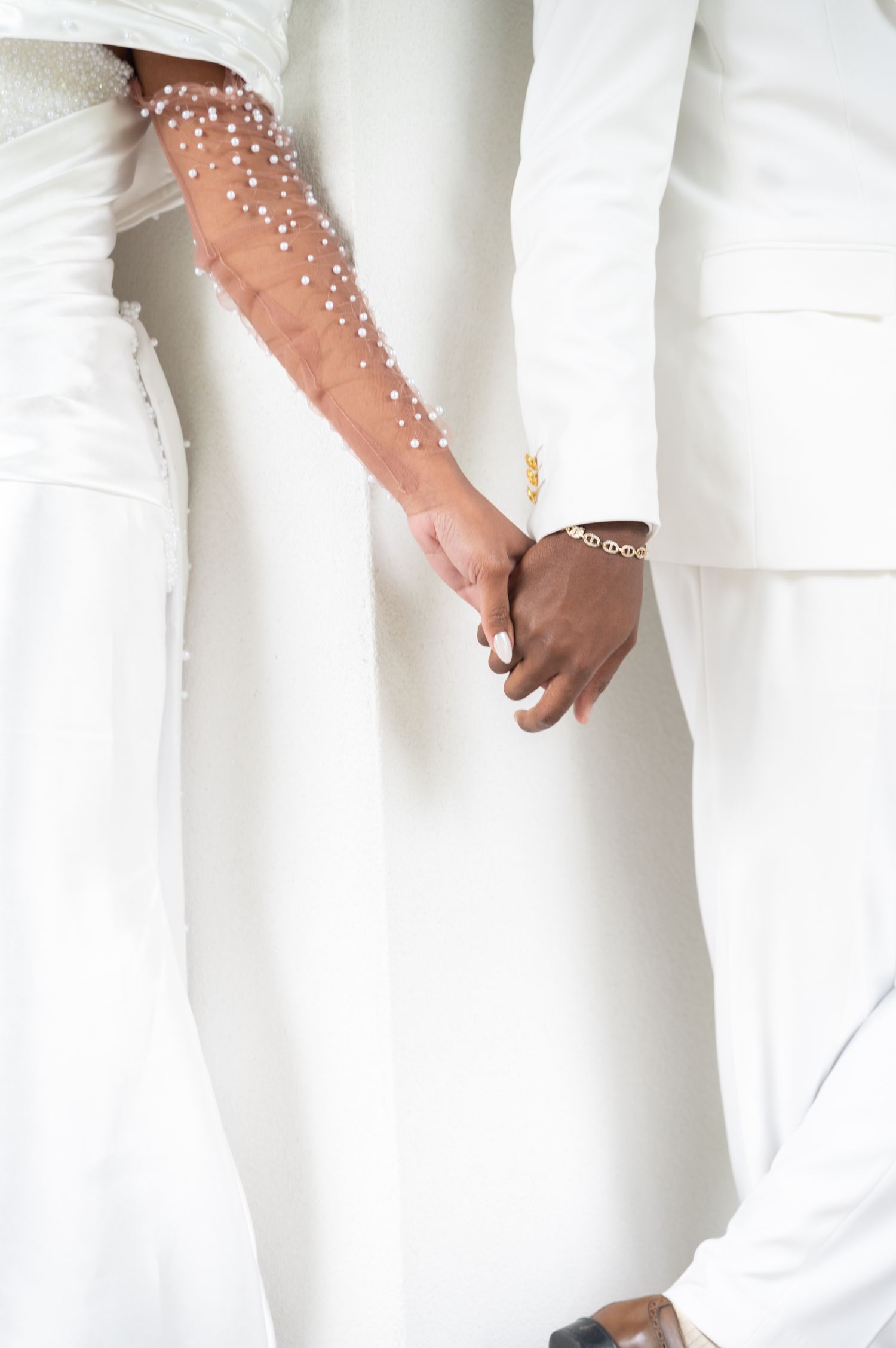 Bride and groom in white attire hold hands, showcasing pearl-embellished bridal sleeve.
