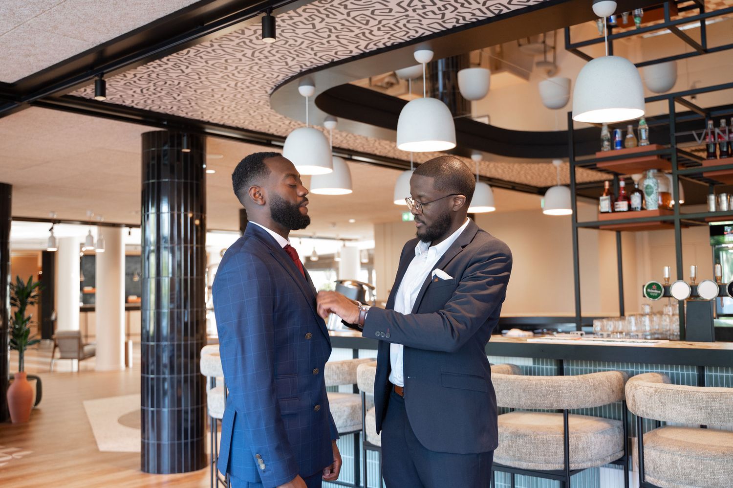 Two Black men in blue suits stand face to face in a stylish modern bar interior during wedding prep.