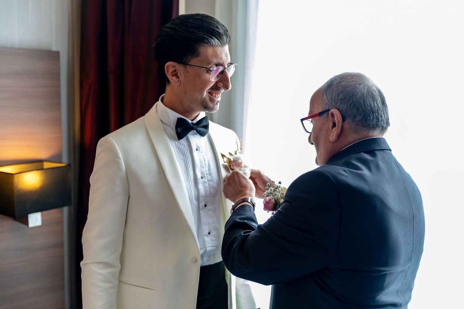 Man in white tuxedo gets boutonniere pinned by older gentleman in hotel room before wedding.