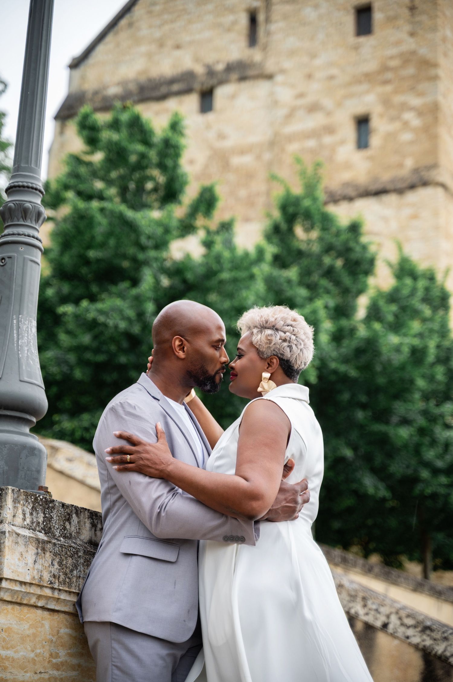 Black couple embracing and kissing near a stone bridge with historic building in background.