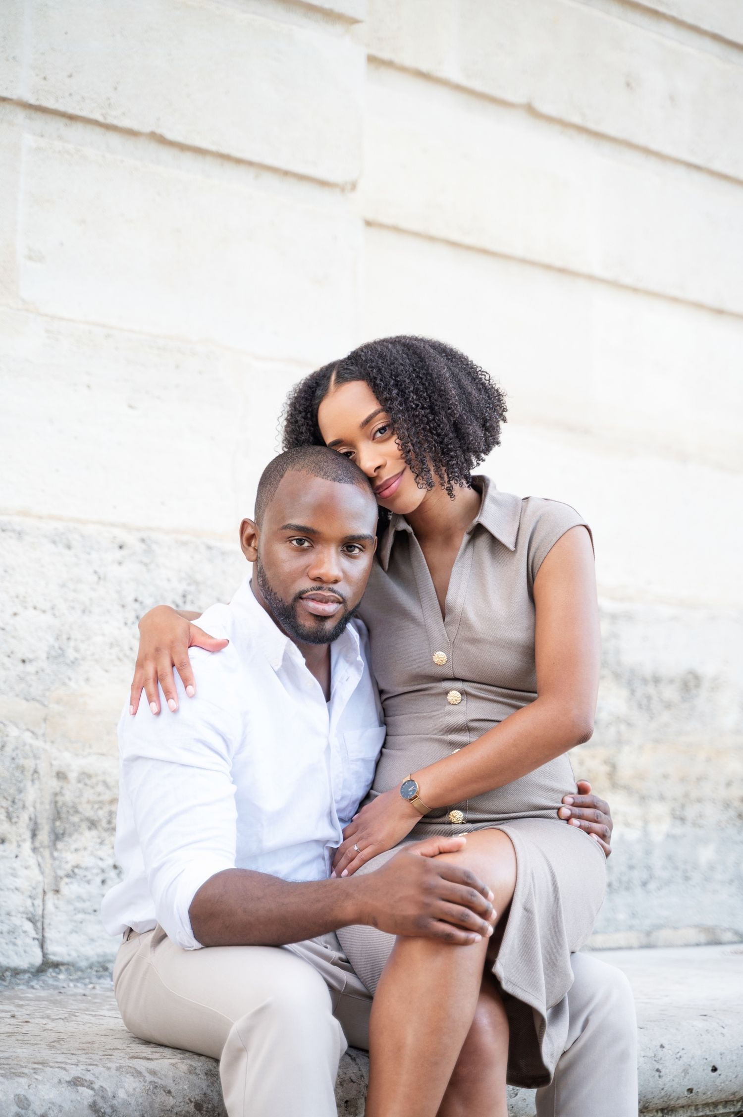 A couple poses on stone steps, the woman in a taupe dress resting her arms around the mans shoulders.