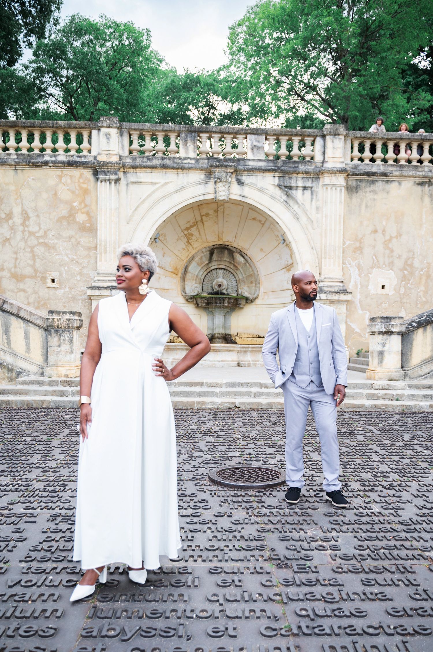 Black couple standing on cobblestone plaza with Latin text, woman in white jumpsuit, man in gray suit.