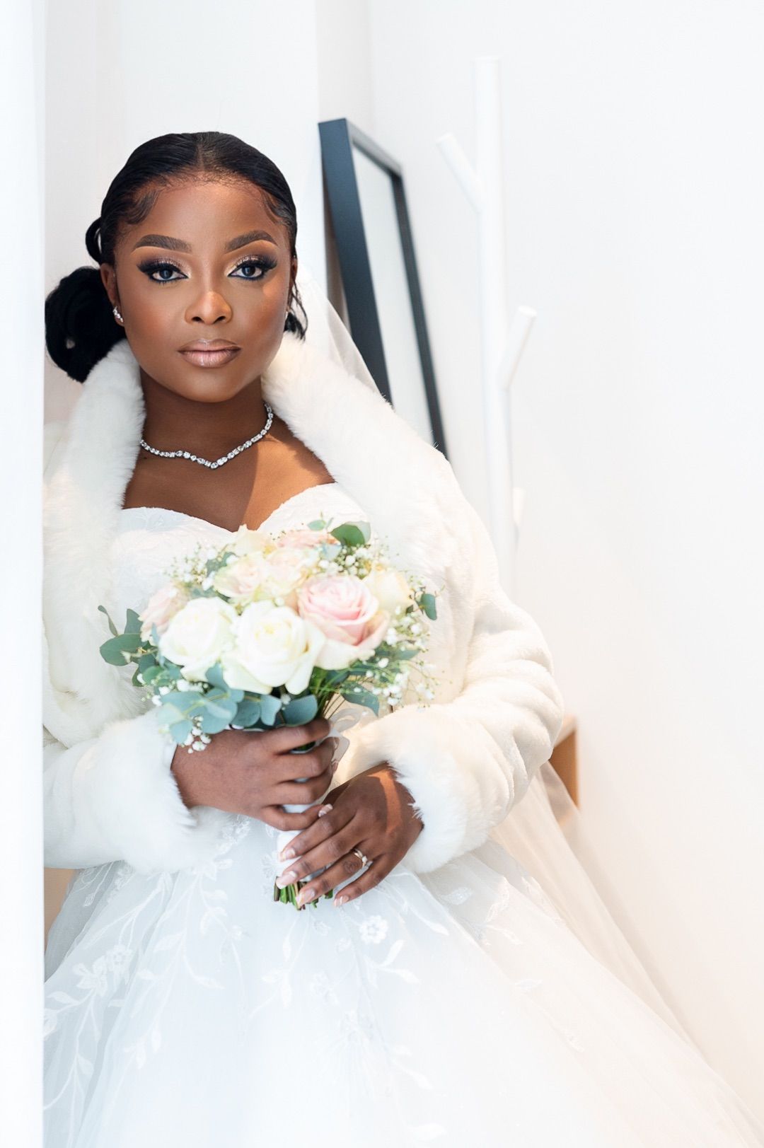 Black bride in white strapless ball gown, fur wrap, and veil holding soft pink and white bouquet.