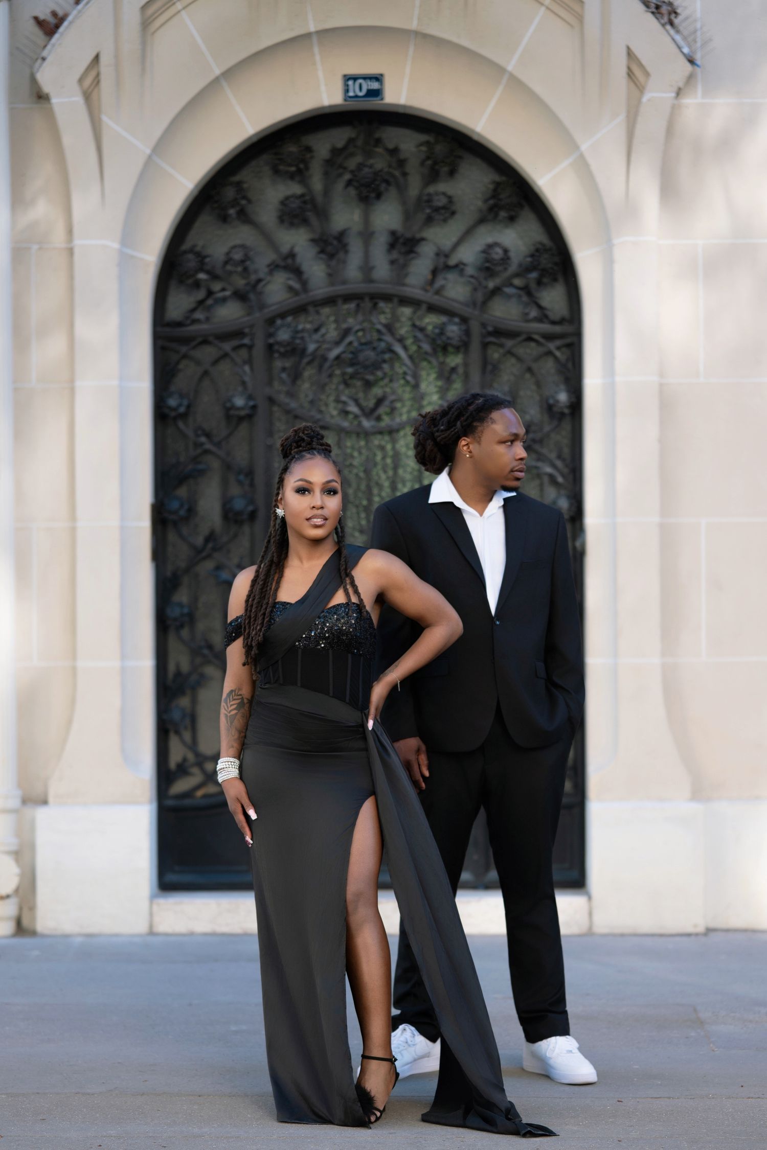 A stylish couple poses in front of an ornate arched doorway, wearing elegant black formal attire.