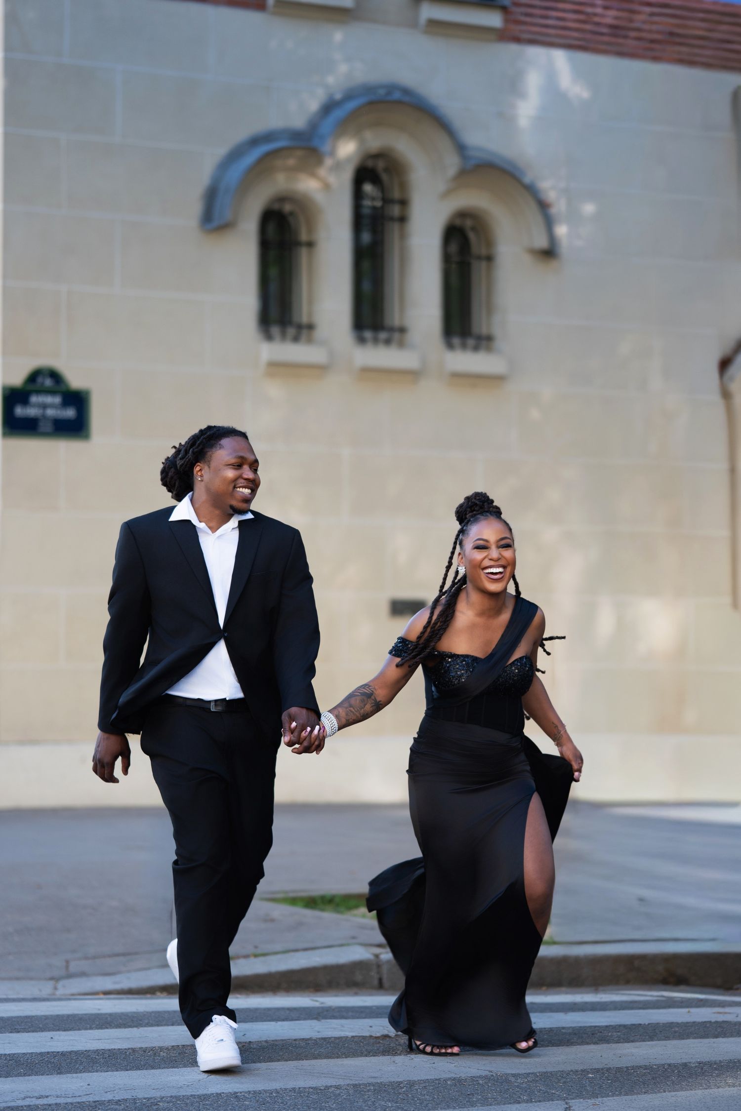 Elegant Black couple in matching black outfits holding hands and laughing while crossing a street in Paris.