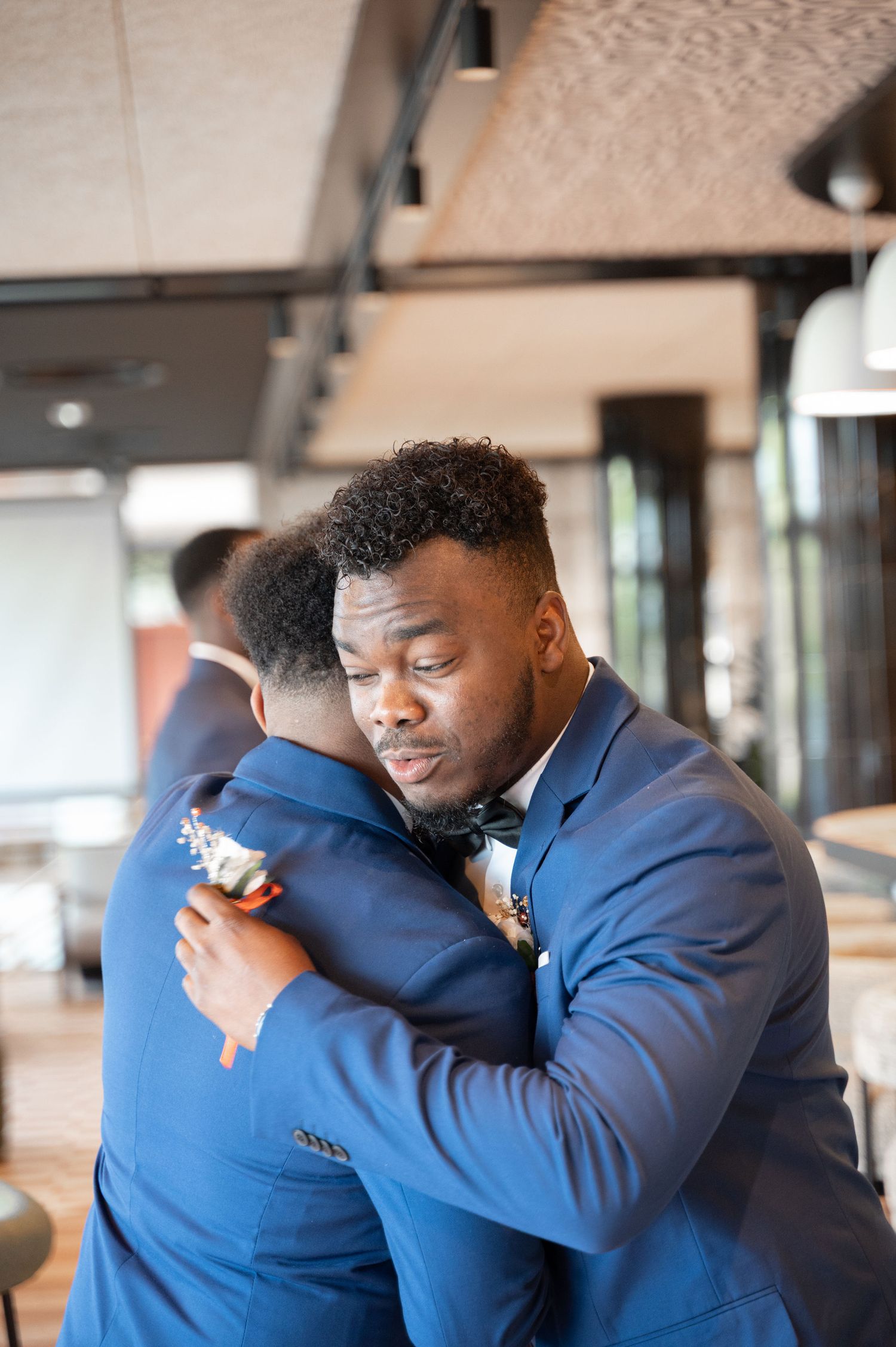 Two Black men in blue suits sharing an emotional hug at a wedding celebration indoors.