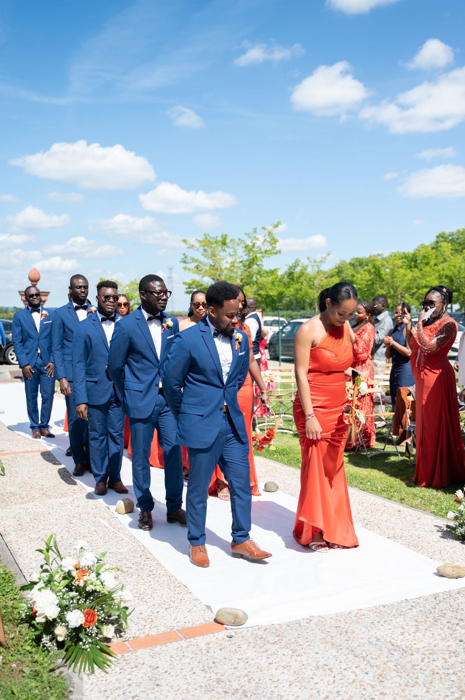 Wedding party procession with groomsmen in navy suits and bridesmaids in orange dresses on white aisle.