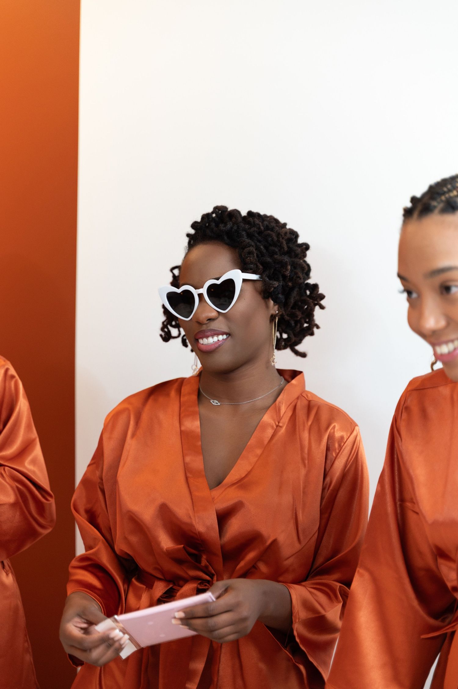 Bridesmaids wearing matching burnt orange satin robes, one smiling with white heart-shaped sunglasses holding phone.