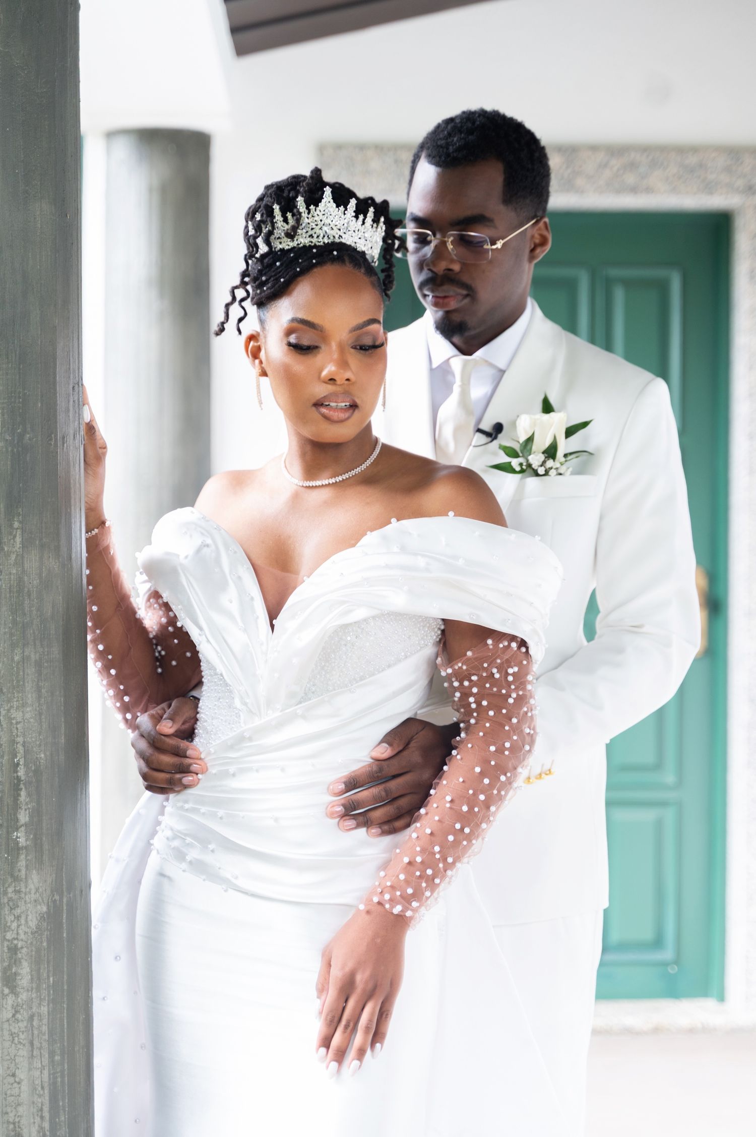 Groom in white suit tenderly embraces bride in pearl off-shoulder gown and diamond crown.