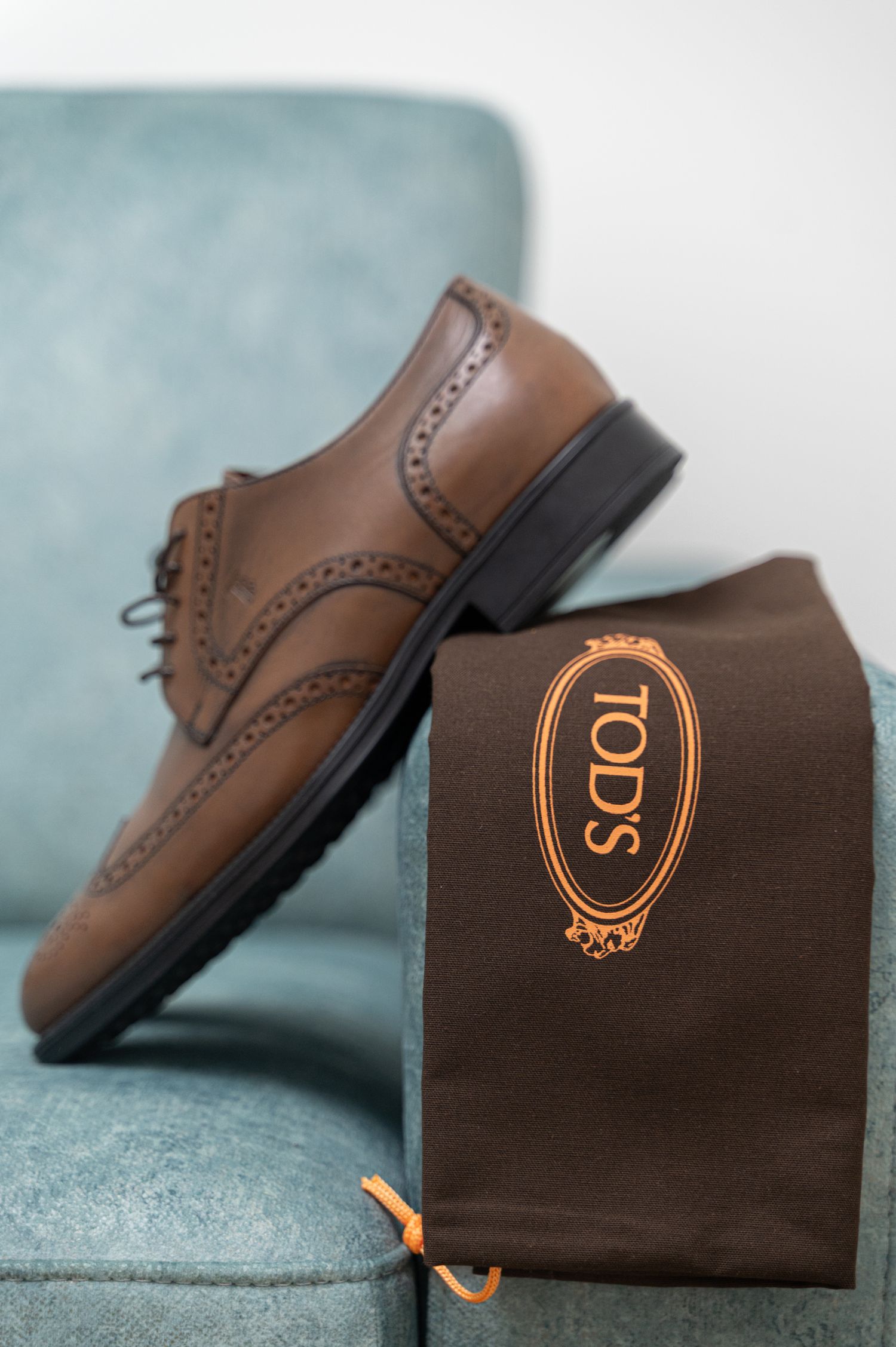 Brown leather Tod's brogue dress shoes displayed on teal chair with branded dust bag.