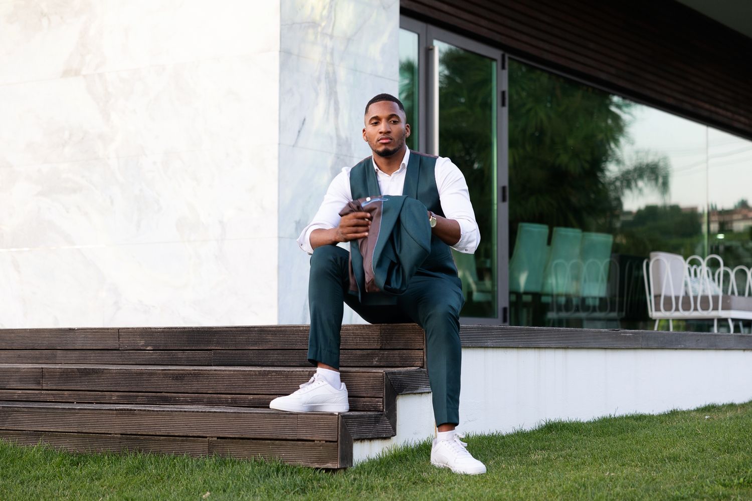 A Black man in a teal suit sits on wooden steps outside a modern building holding a smartphone.
