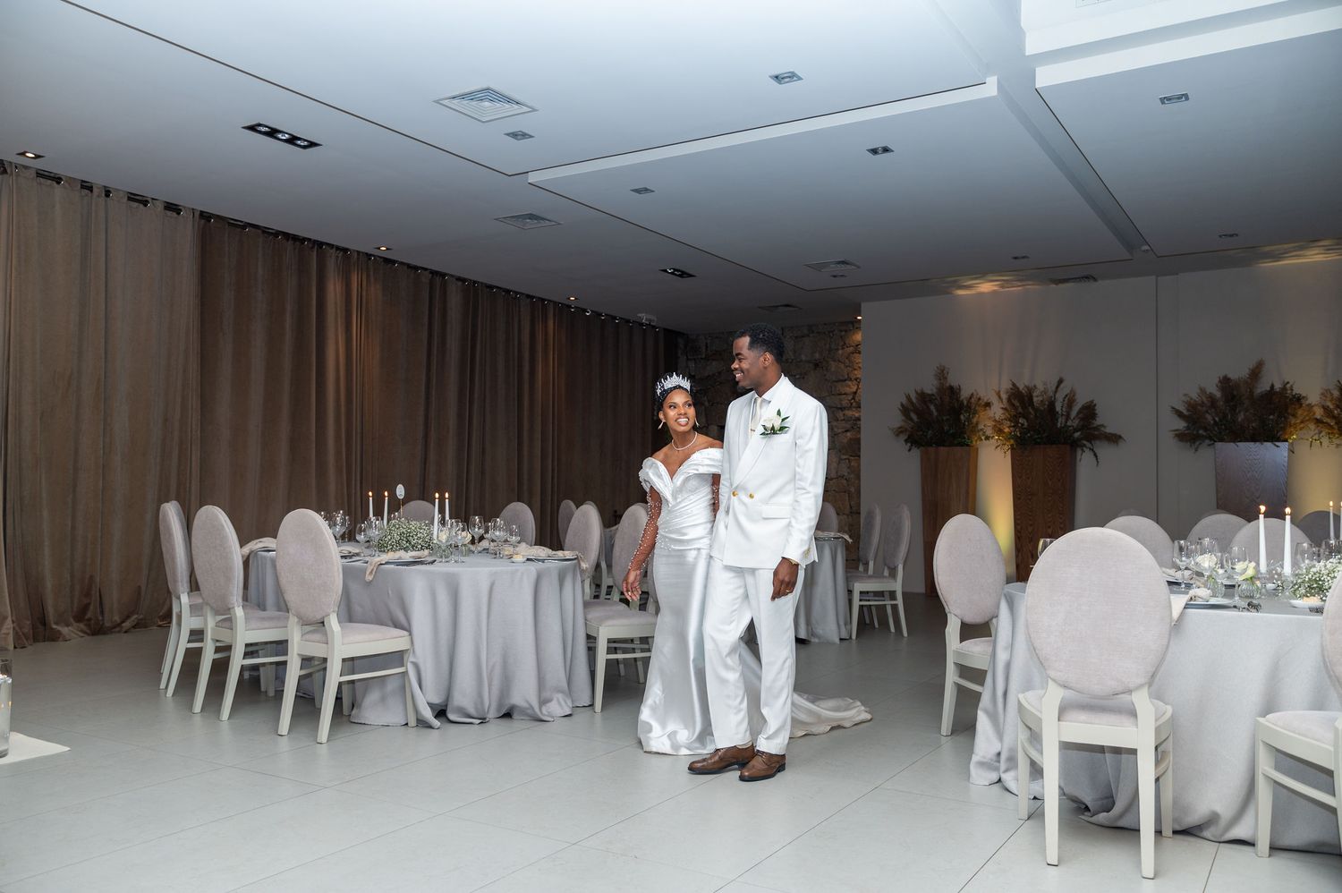 A Black bride and groom in white attire stand in a beautifully decorated wedding reception venue.