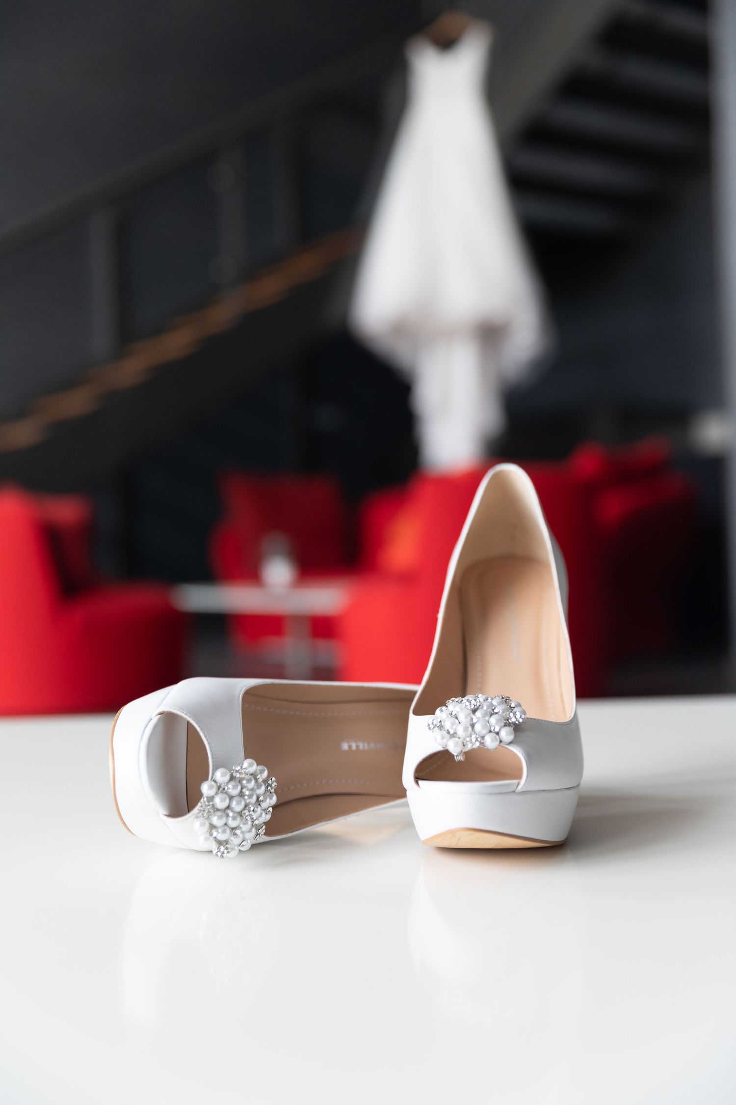 Elegant white bridal heels with crystal embellishments displayed on a table with a wedding dress in the background.