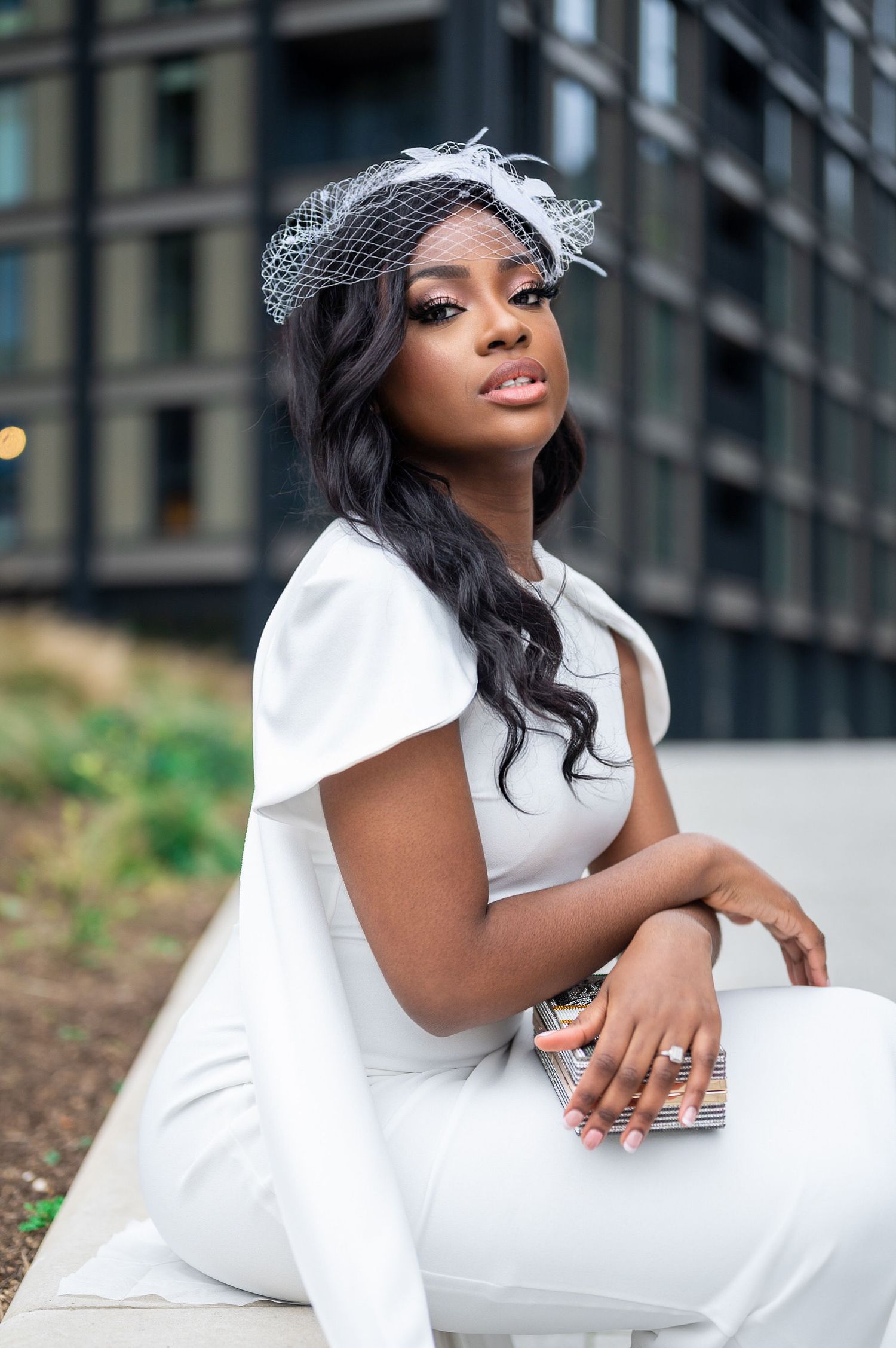 An elegant Black bride poses in a white jumpsuit and birdcage fascinator, seated outdoors near a building.
