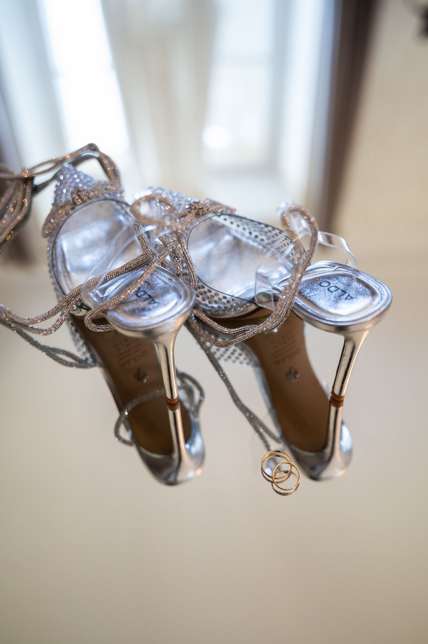 Silver strappy wedding heels displayed upside down with gold wedding rings placed between them.