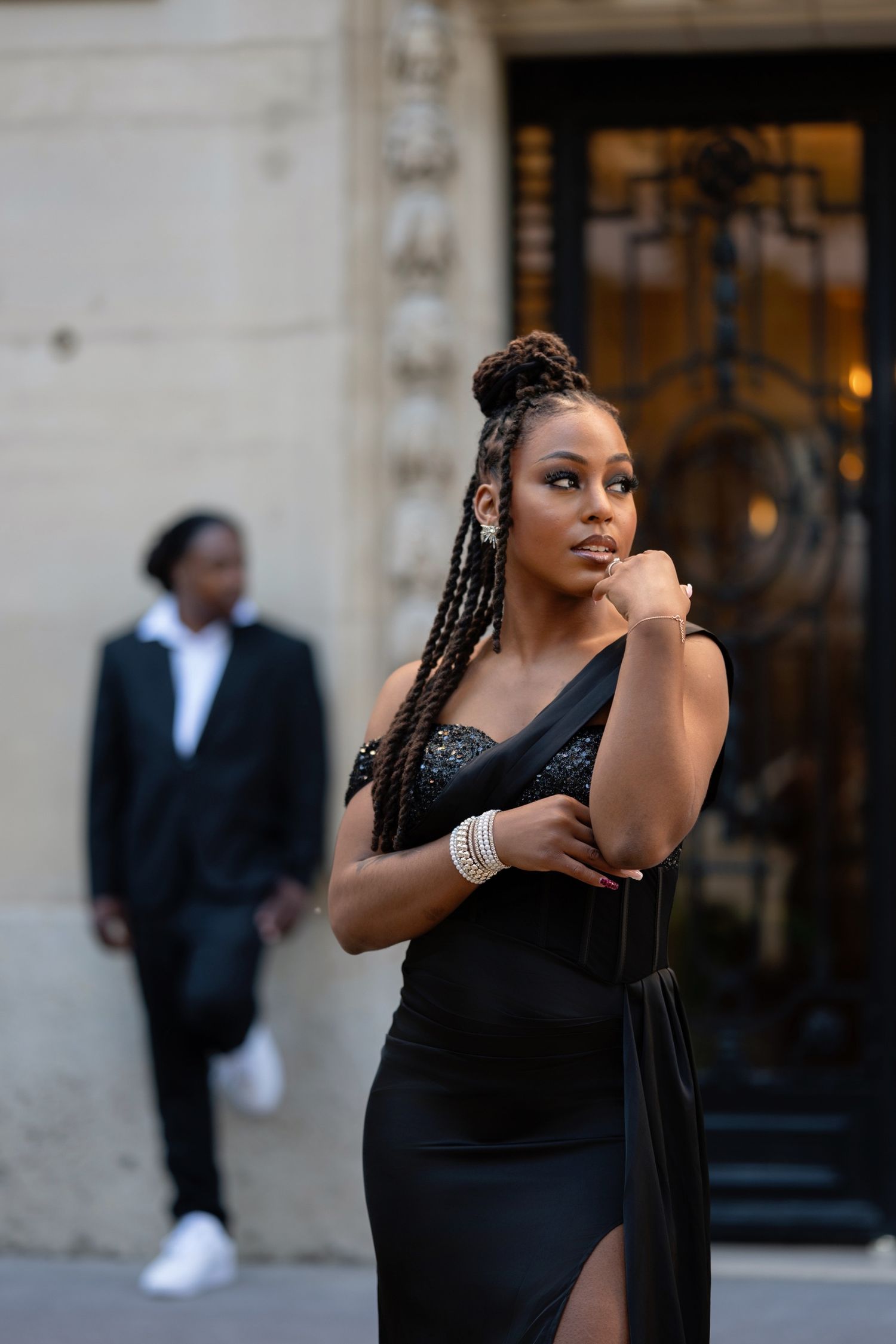 A woman in a black slit gown with braids poses confidently outdoors, with a man in a suit visible in the background.