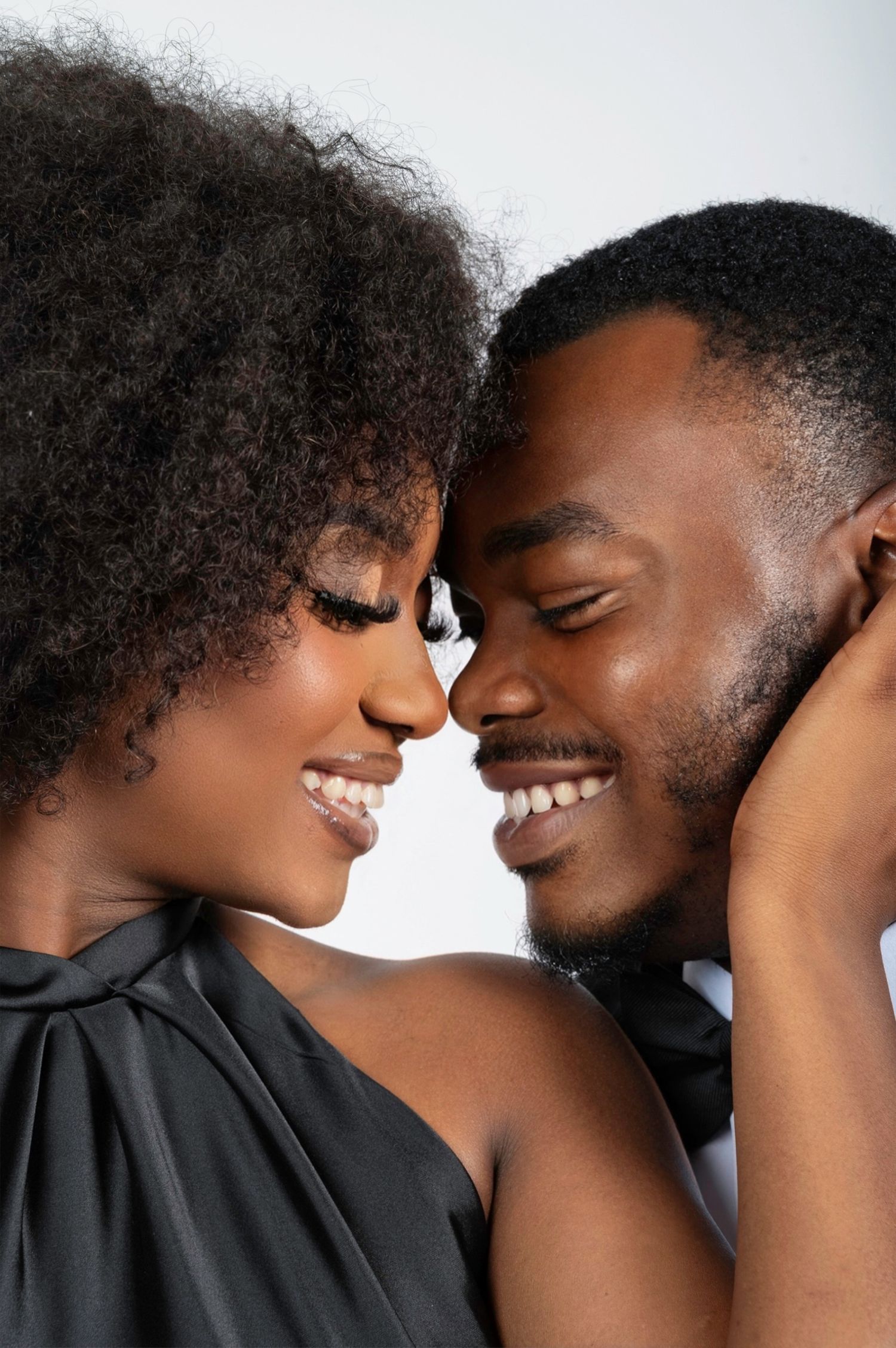 Smiling Black couple in elegant black attire sharing an intimate moment, foreheads touching against white background.
