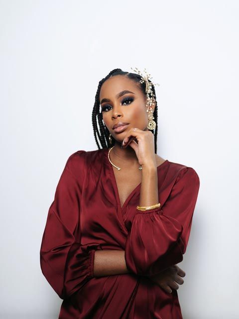 Elegant woman in burgundy satin wrap dress with box braids and gold jewelry posing confidently.