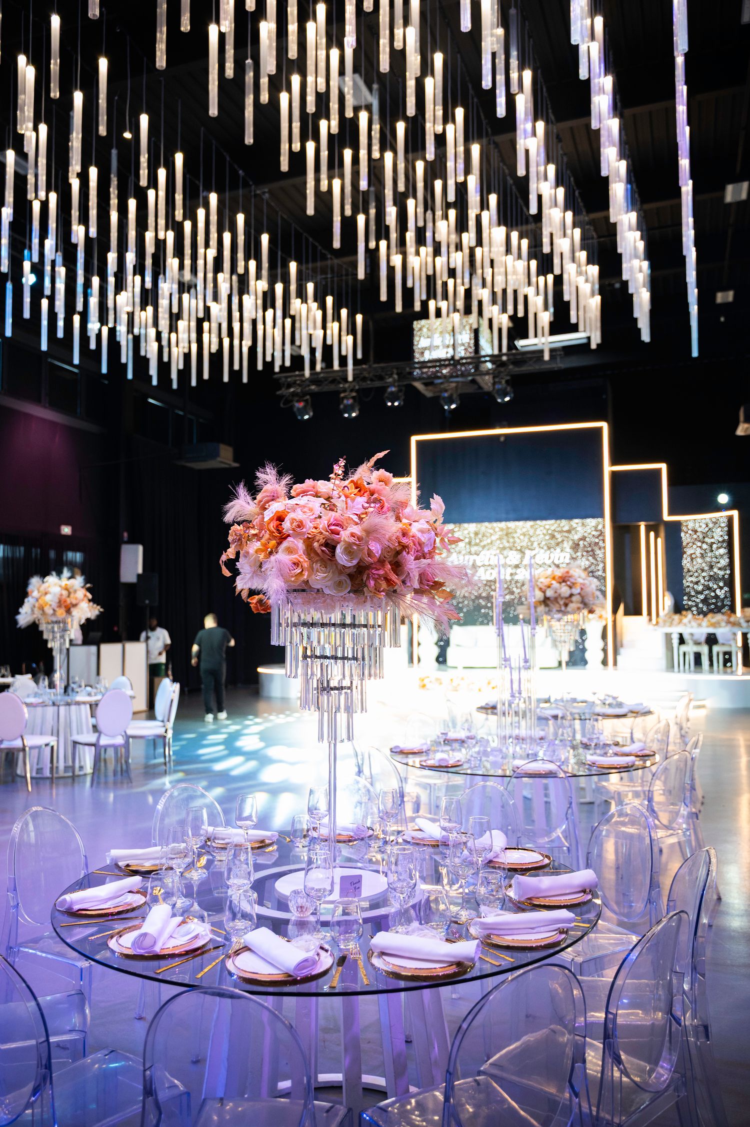 Luxurious wedding reception table with tall pink floral centerpiece, ghost chairs, and dramatic ceiling lights.