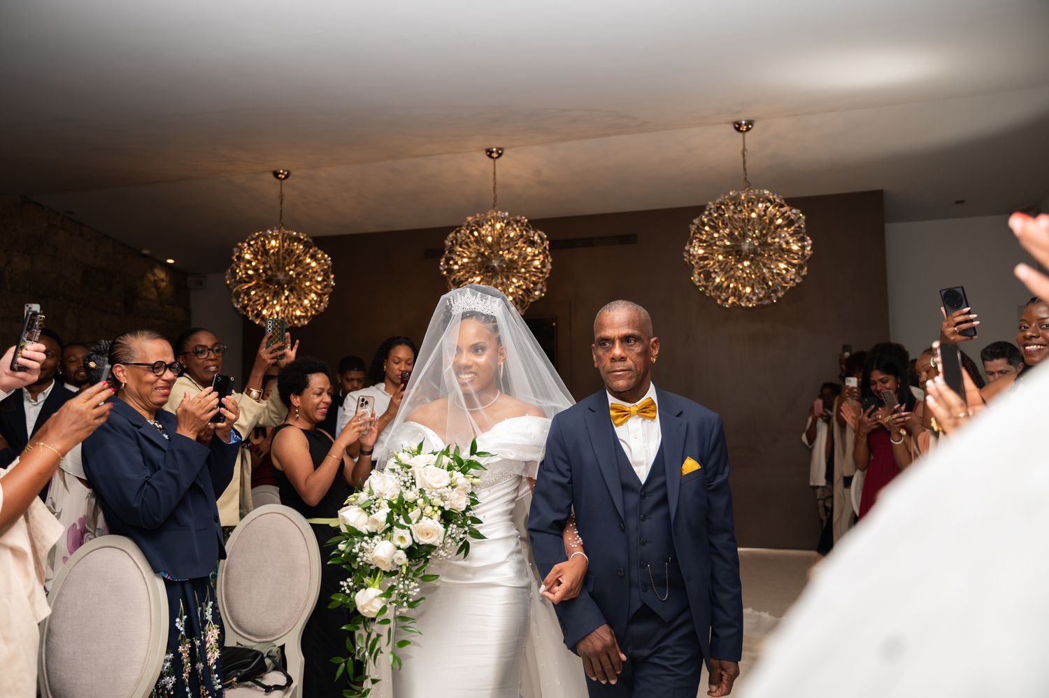 Bride in white gown and veil walks down the aisle escorted by her father at an elegant wedding.