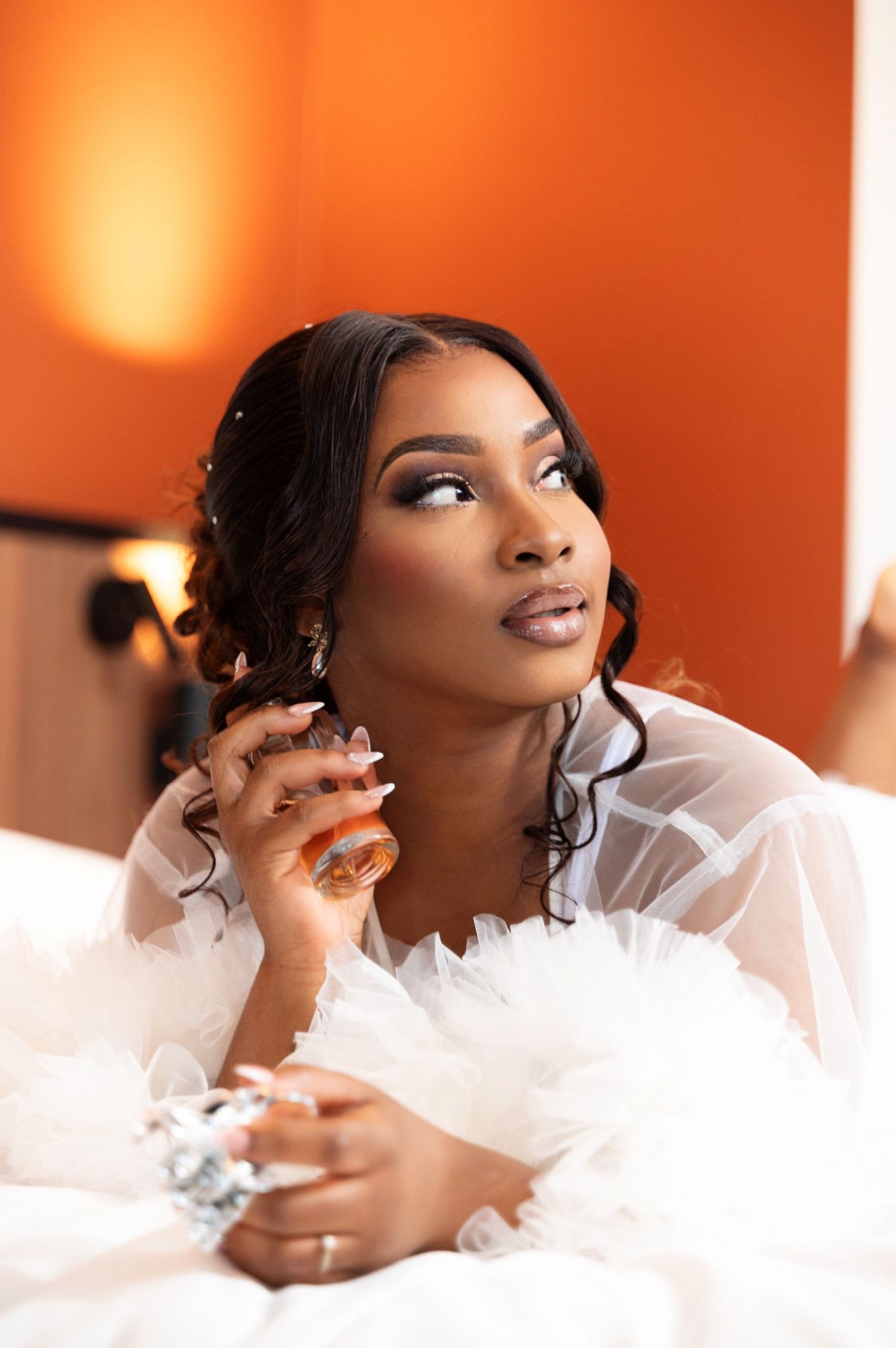 Stunning bride in white feathered robe lying on a bed gazing upward against a bold orange wall.