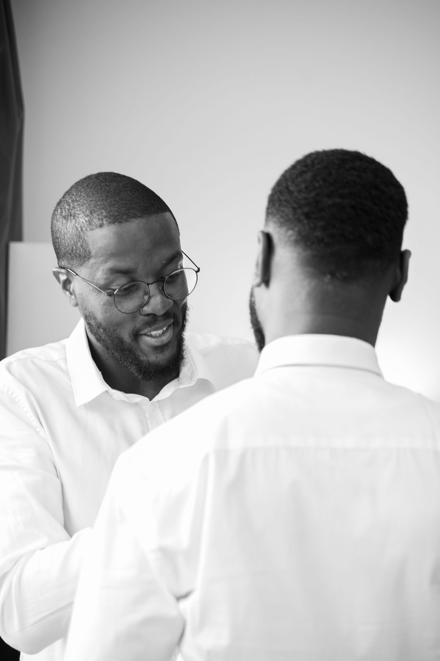Two well-dressed Black men in white shirts share a warm moment as one adjusts the other's clothing.