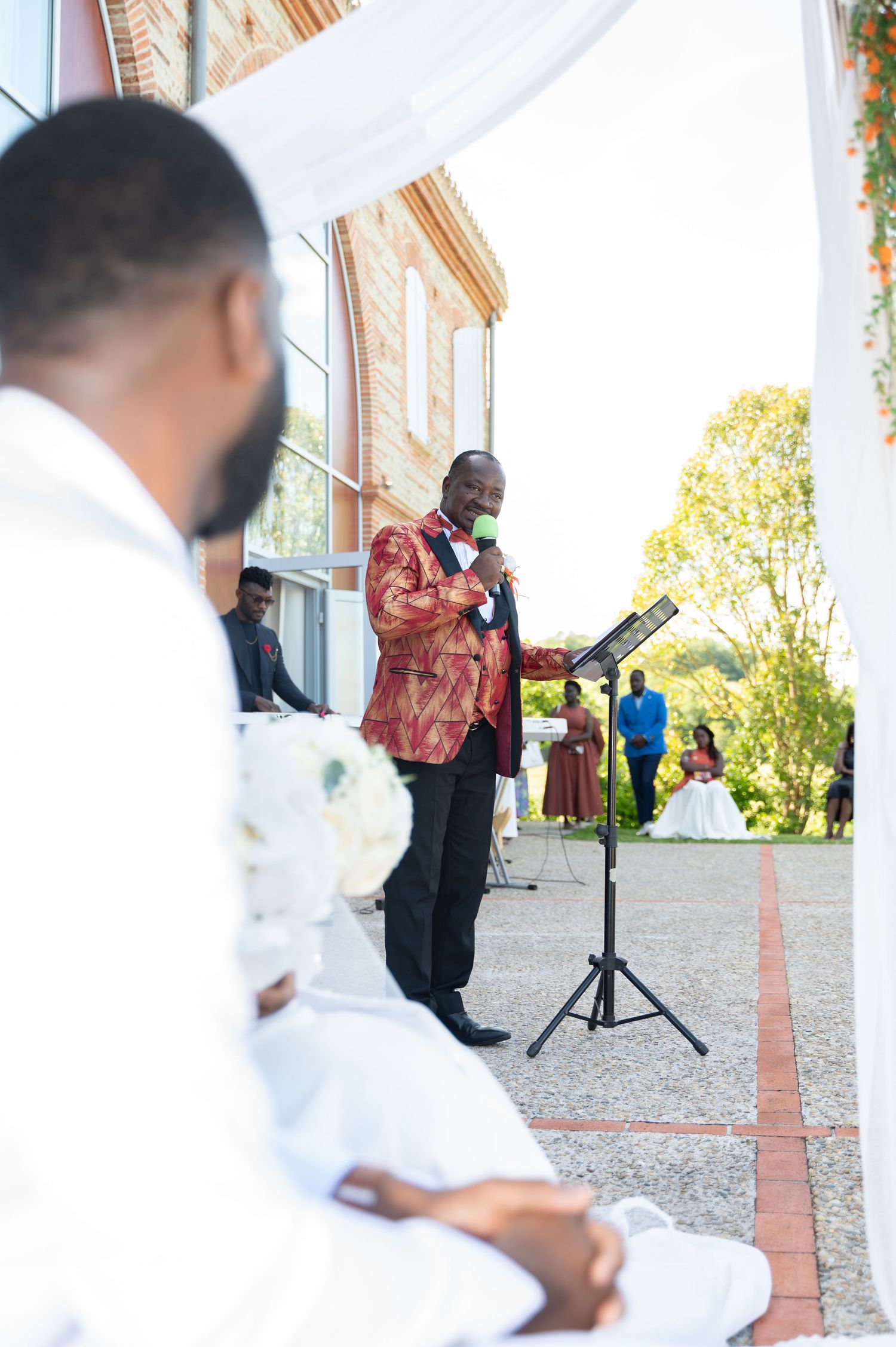 Wedding officiant in colorful African attire speaks at outdoor ceremony podium.