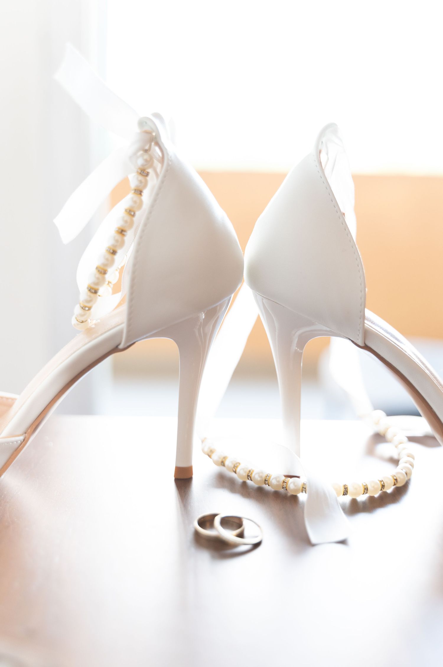 White bridal heels with pearl necklace and wedding rings on wooden surface.
