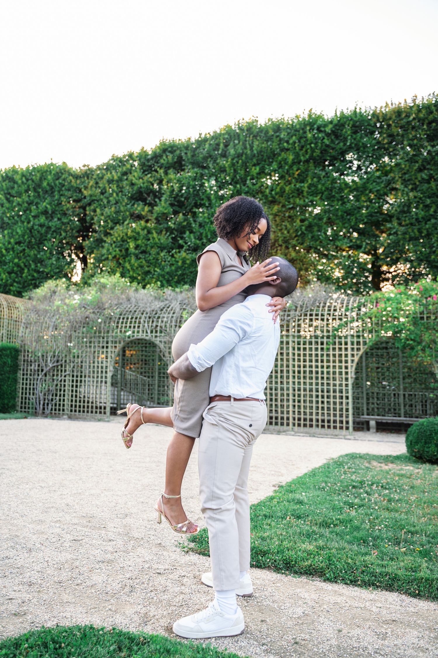 A joyful Black man lifts his smiling partner in a taupe dress in a beautifully manicured garden setting.