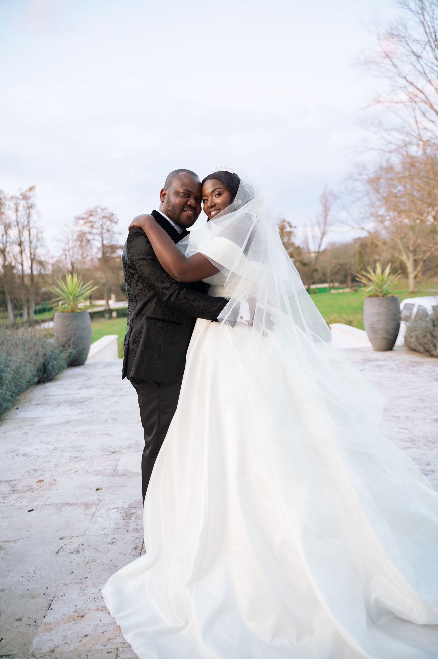 Bride in flowing white ball gown with veil embracing groom in black suit in garden setting.