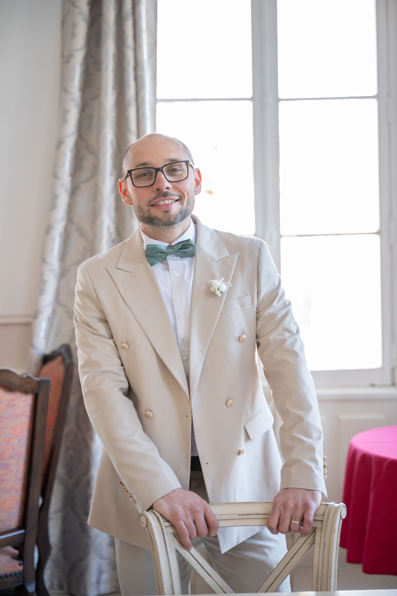 Elegant groom in cream double-breasted suit with green bow tie and glasses posing by chair near window.