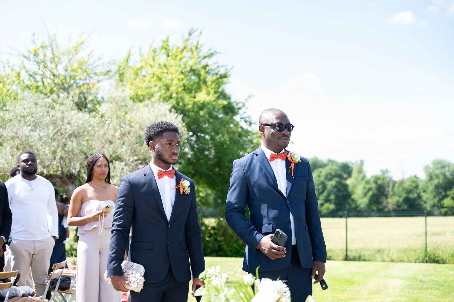 Two groomsmen in navy suits with orange bow ties walk down the aisle at outdoor wedding.