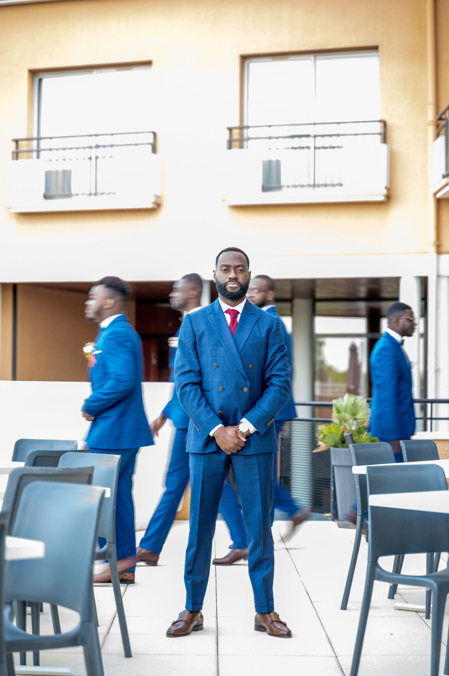 Elegant Black groom in navy double-breasted suit stands poised on outdoor patio with blurred groomsmen behind.