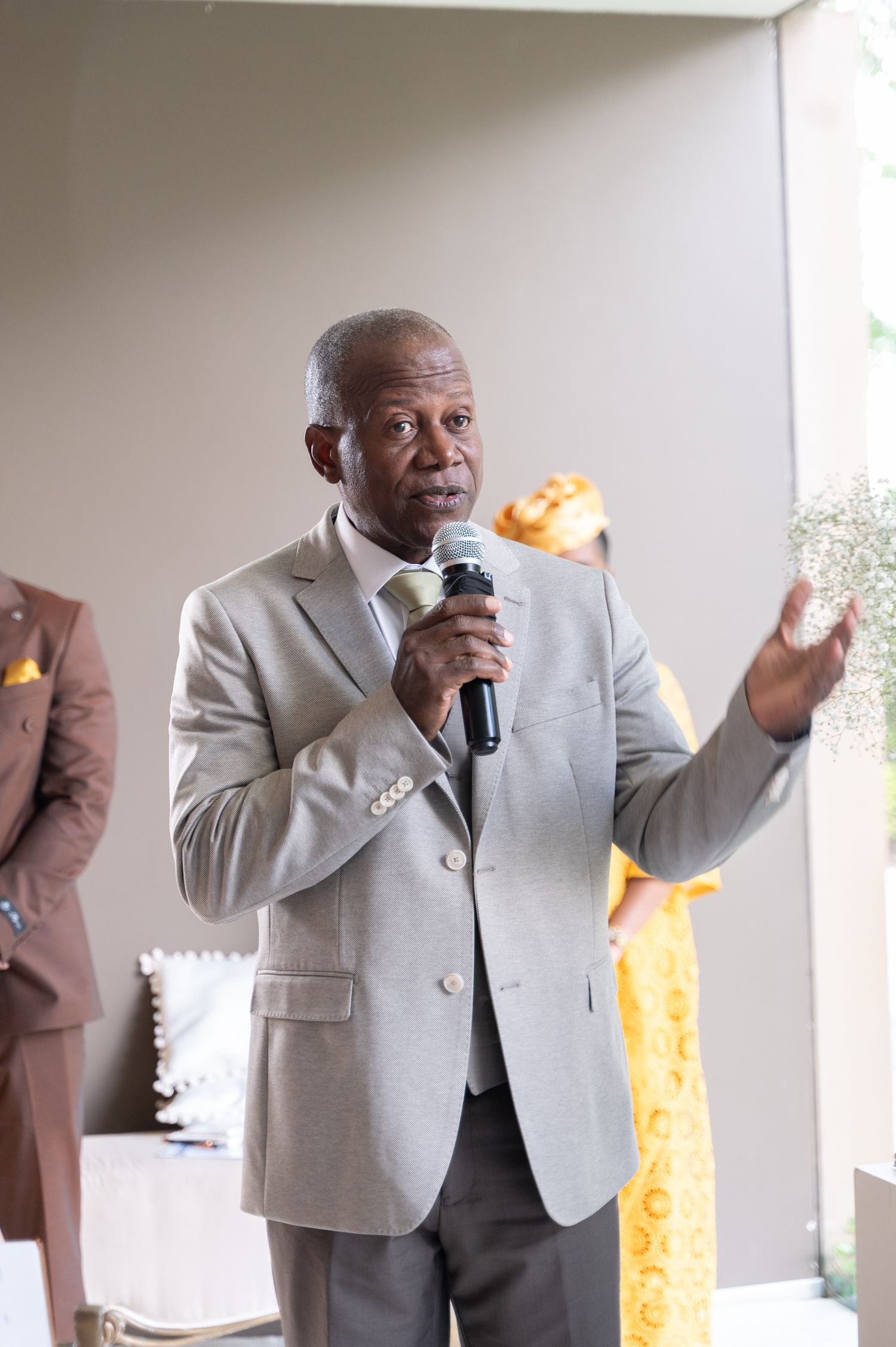Elderly man in gray suit giving speech with microphone at wedding reception.