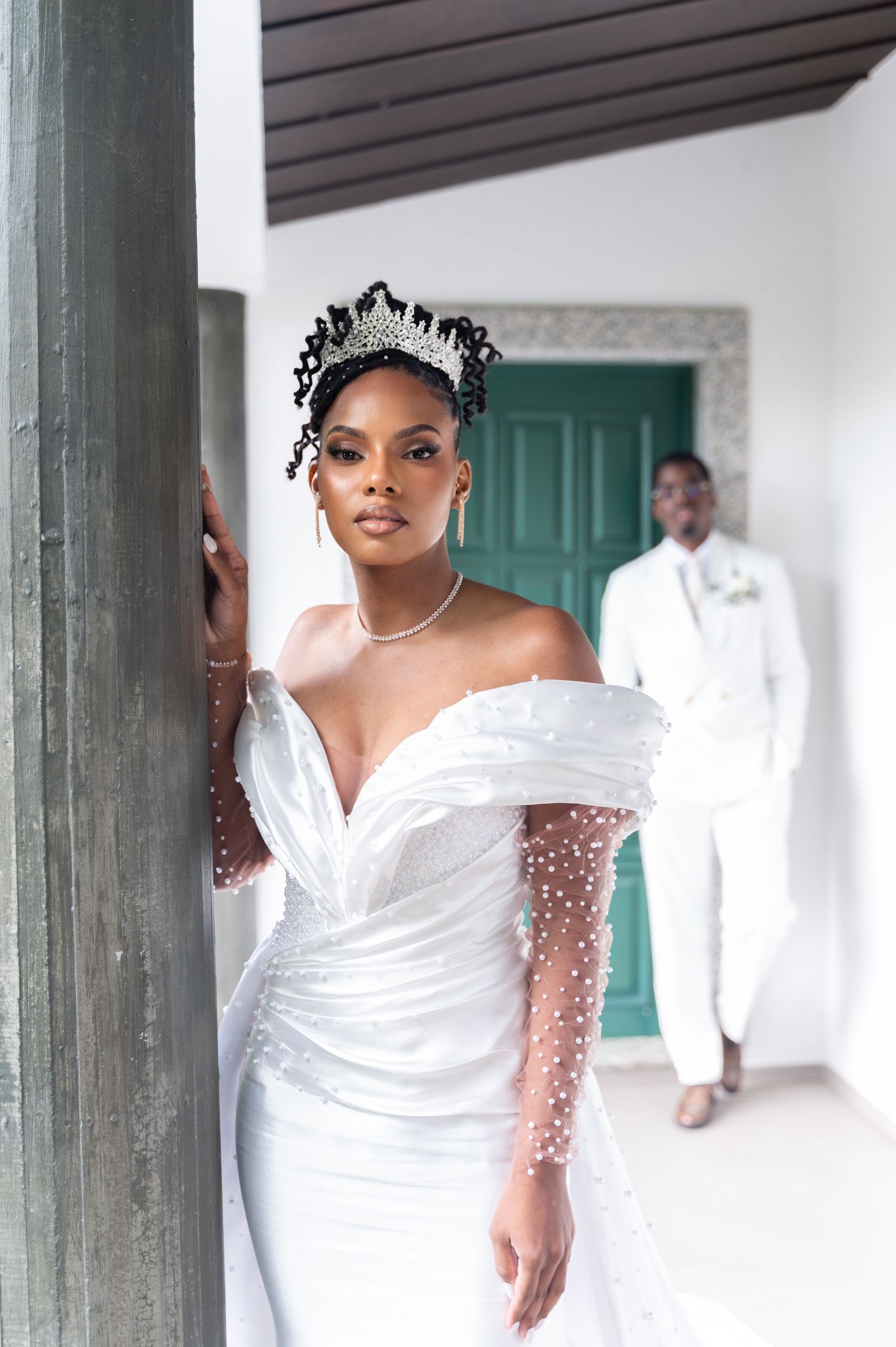 Beautiful Black bride in off-shoulder pearl wedding dress and diamond tiara with groom in background.