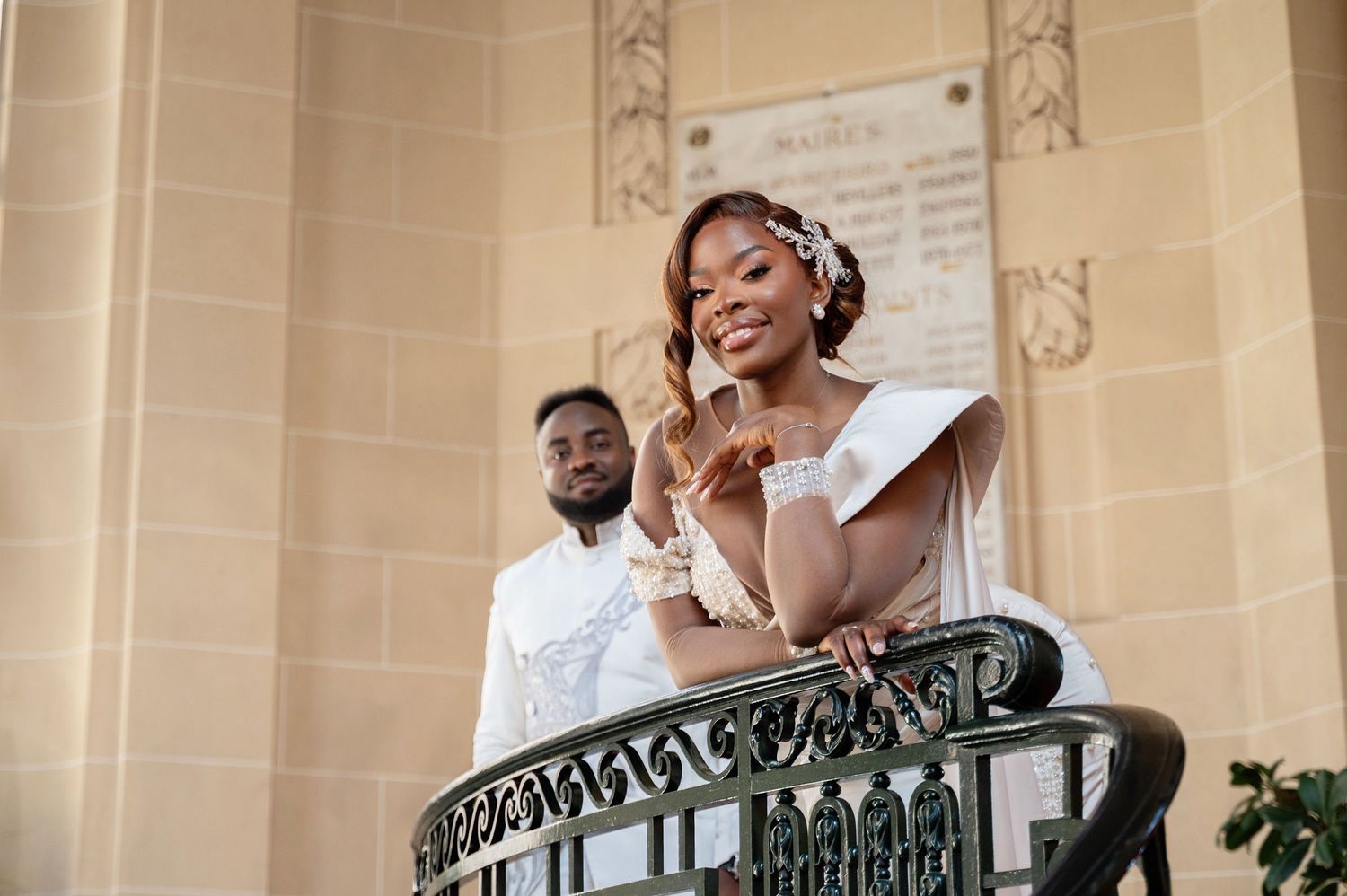 Smiling bride in white off-shoulder gown stands on ornate balcony with groom behind her.