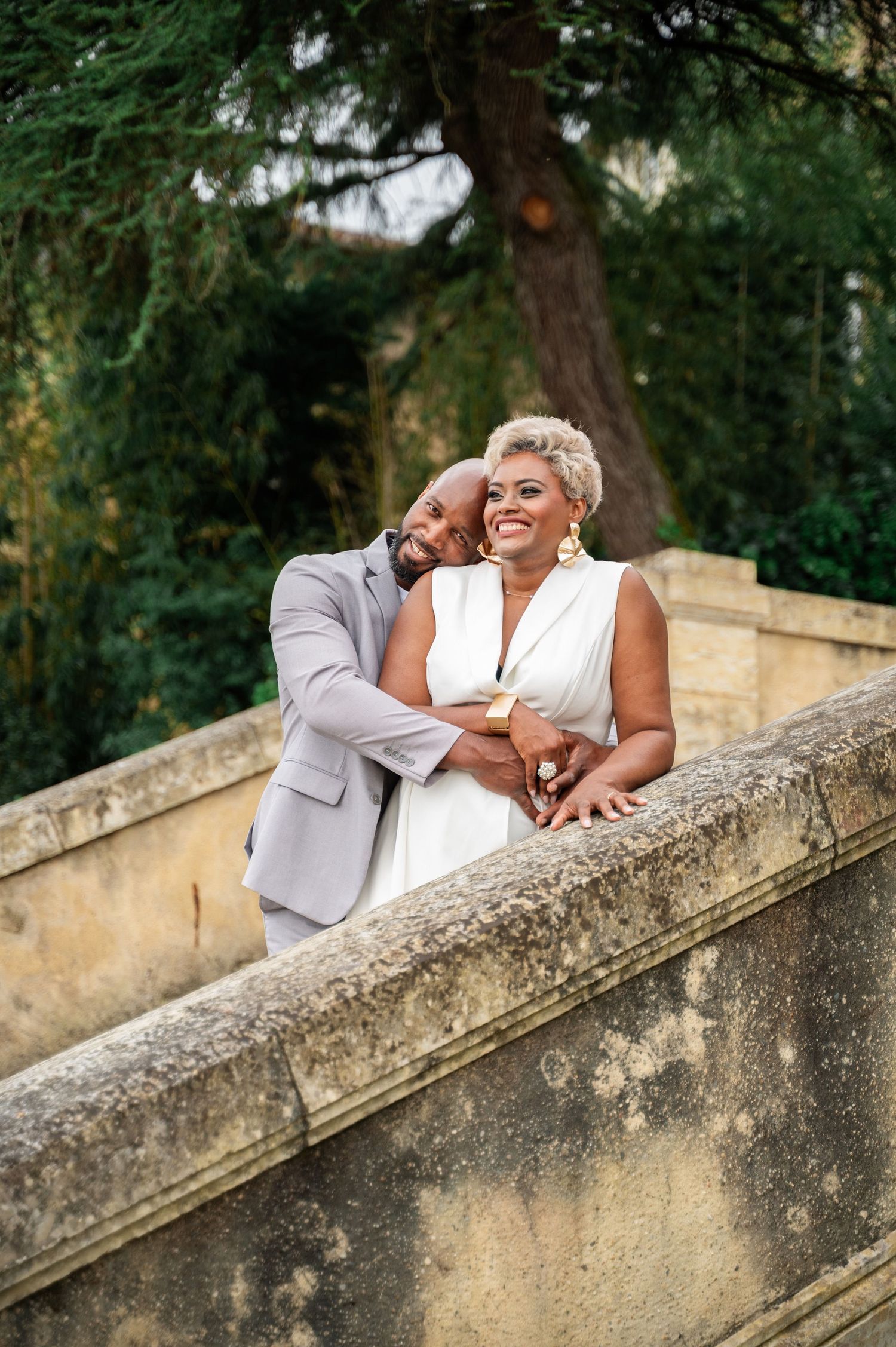 Romantic Black couple embracing on stone balustrade, man in gray suit nuzzling woman in white dress.